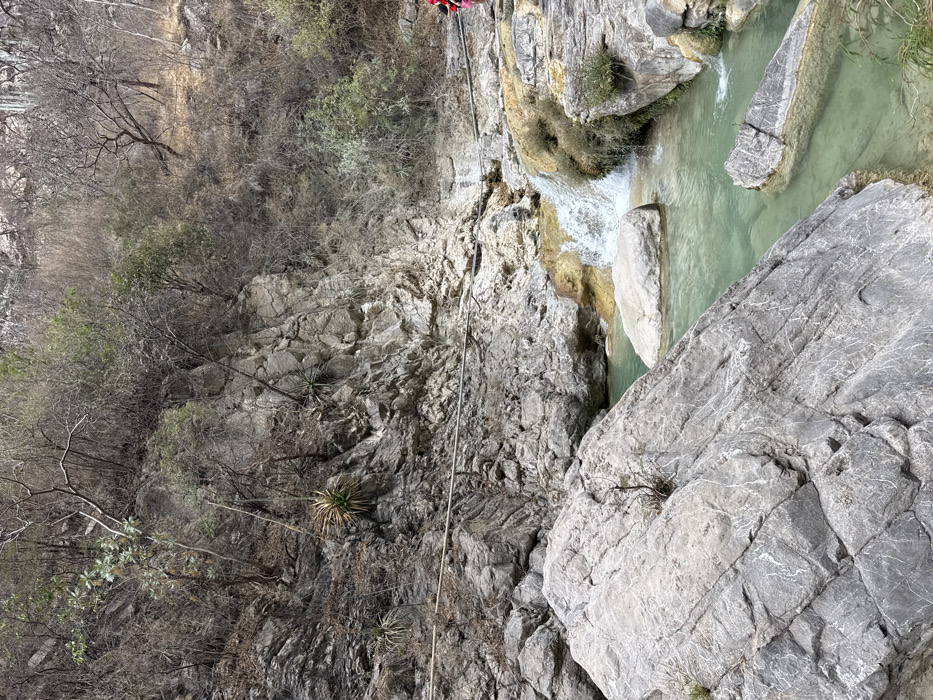 The image depicts a natural canyon landscape in San Miguel Atlapulco, Mexico, characterized by a stream, waterfall, and rocky terrain under an overcast sky. In the foreground, large, grey, angular rock formations with visible striations dominate the lower left. A clear, greenish-blue stream flows from right to left, encompassing these rocks and several submerged boulders. In the midground, a small waterfall cascades approximately one meter into the stream from a higher, yellowish-tan rock formation, indicating mineral deposits. A horizontal line, resembling a cable or rope, spans across the scene, affixed to the steep rock face above the water. A person, partially visible on the far right, wears a bright red top and appears to be standing on the stream bank. The background consists of a steep, rugged cliff face of grey rock, densely covered with sparse, dry, deciduous vegetation and several agave-like succulent plants. The overall environment suggests a dry or semi-arid climate.