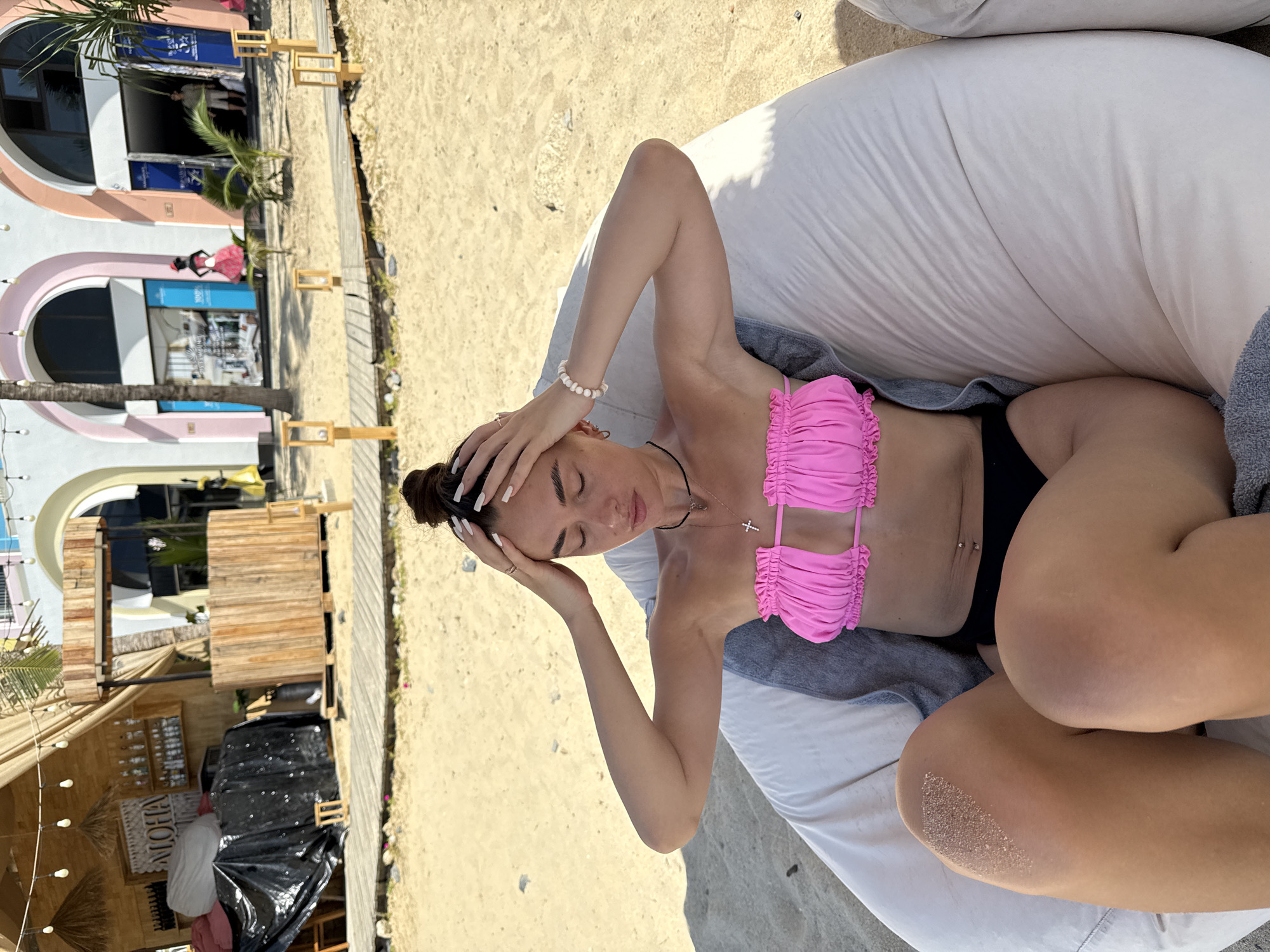 A young woman with dark hair pulled up in a bun is reclining on a large white beanbag chair on a sandy beach. She is wearing a bright pink ruched bikini top and black bikini bottoms. Her eyes are closed, and her hands are resting on her head, suggesting relaxation or a moment of quiet contemplation. She is wearing a beaded bracelet on her left wrist and a delicate necklace with a cross pendant. A grey towel is draped over the beanbag chair, partially covering her legs. The background shows a sandy beach with wooden walkways and structures, suggesting a beach resort or a similar establishment. In the distance, a building with arched windows and pink and white accents is visible, along with palm trees. The scene appears to be set during the daytime, with bright sunlight casting shadows. The presence of a "Aloha" sign suggests a tropical or beach-themed location. The overall mood conveyed is one of leisure and relaxation in a sunny, beachside setting.