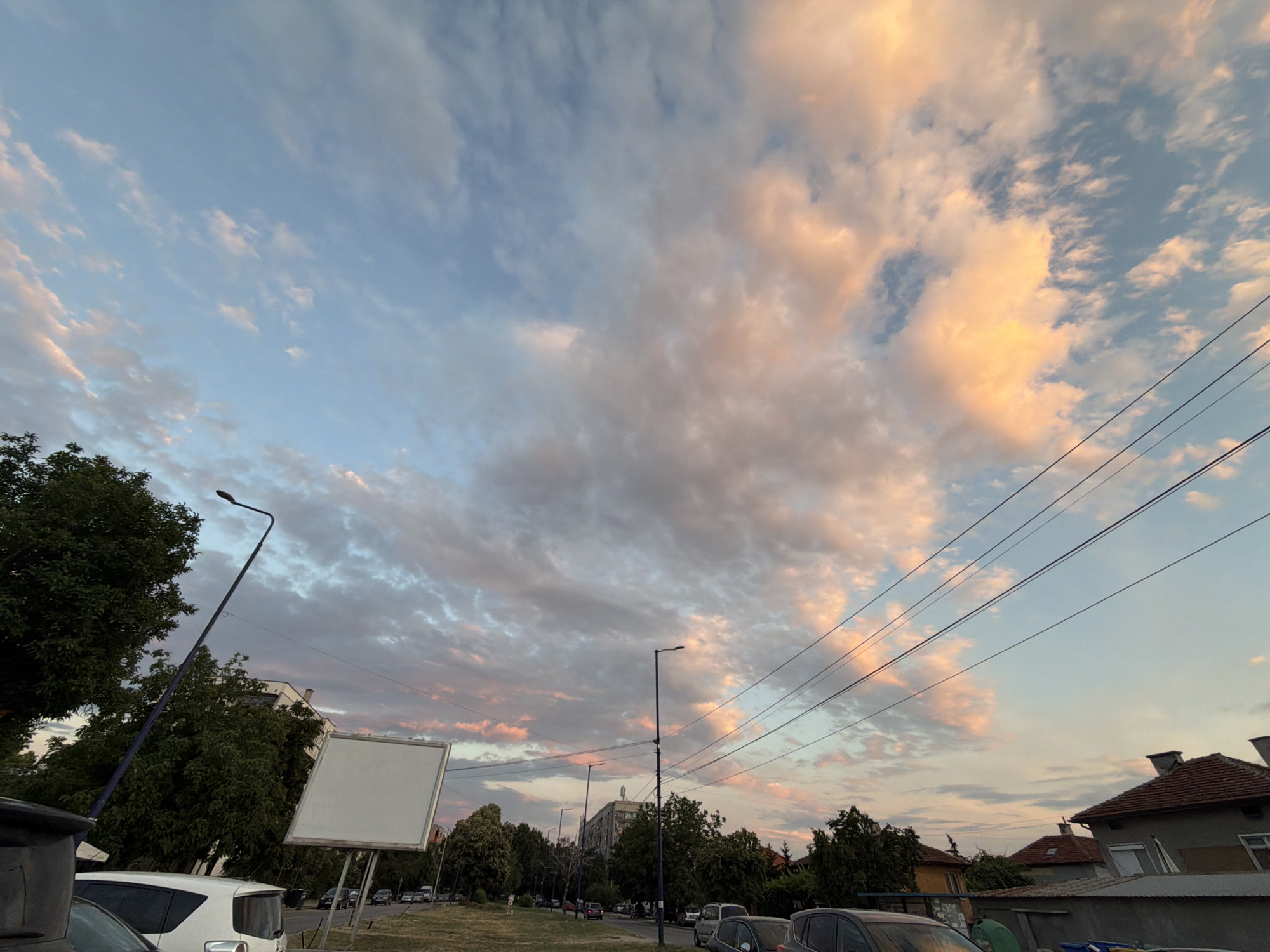 The image depicts a suburban street scene at dusk, with a sky filled with pink and orange-tinged clouds. The foreground shows the tops of several cars, one white van, and a black SUV. To the left, a tall, dark lamp post with a blue pole stands behind a dense green tree. In the mid-ground, a large, blank white billboard is angled towards the viewer, its support poles visible. Further back, more trees line a grassy median strip alongside a street with cars driving and parked. Several other street lamps are visible along the road. Buildings with tiled roofs and windows are scattered among the trees on the right side of the image. Overhead, multiple dark power lines crisscross the sky.