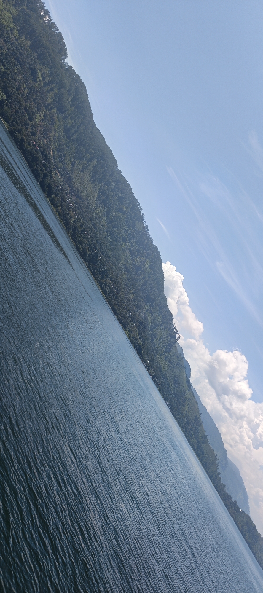 The image shows a landscape view of a body of water with a forested mountain slope. The water is a deep blue with ripples, suggesting it is a lake or a calm sea. The mountain is covered in dense green vegetation and is illuminated by sunlight. There are some white clouds in the sky, which is a bright blue. The scene is tilted, with the horizon line running diagonally across the image.
This scene is characteristic of the natural beauty found around Tigaras, a sub-district in Simalungun Regency, North Sumatra, Indonesia, which borders Lake Toba. Lake Toba is known for its stunning volcanic caldera, surrounded by lush green hills and mountains. The visual elements in the photograph – the expansive blue water, the forested slopes, and the clear sky with scattered clouds – are consistent with the typical scenery of this region.
