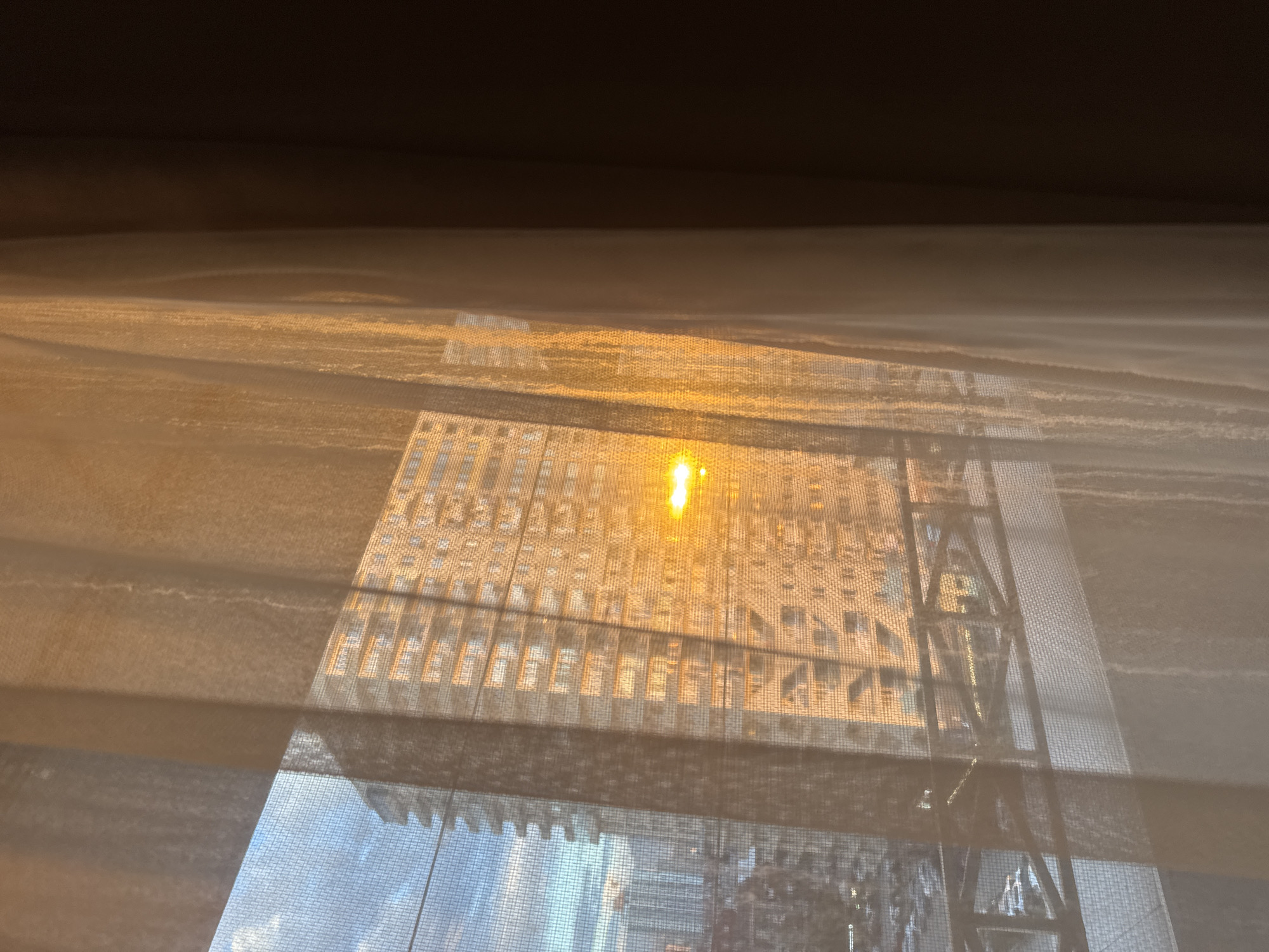 The image shows a view through a window with a translucent curtain or blind partially obscuring the scene. The scene outside appears to be a cityscape, with a large building reflecting sunlight, creating a bright glare. A construction-like structure is visible, possibly scaffolding, partially covering the building. The exact location within the city is unclear, but the scene suggests a dense urban area with tall buildings and construction activity.