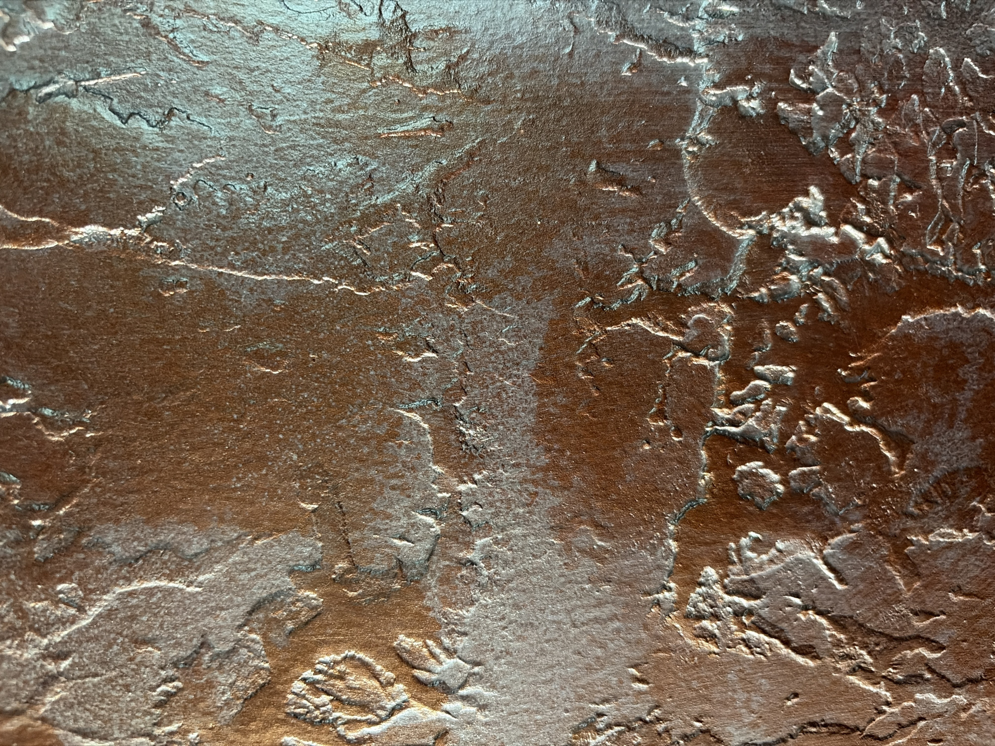 The image displays a close-up view of a textured surface with a metallic sheen. The surface appears to be a rough, uneven material, possibly a wall or a decorative panel, coated in a mottled brown and silver paint. The texture is characterized by raised ridges, valleys, and granular areas. Light reflects off the metallic particles, creating highlights and shadows that emphasize the topographical variations of the surface. The overall impression is one of a weathered or deliberately aged material, with a somewhat industrial or artistic aesthetic. There are no people, identifiable objects, or environmental features visible to suggest a specific location or context within a city.