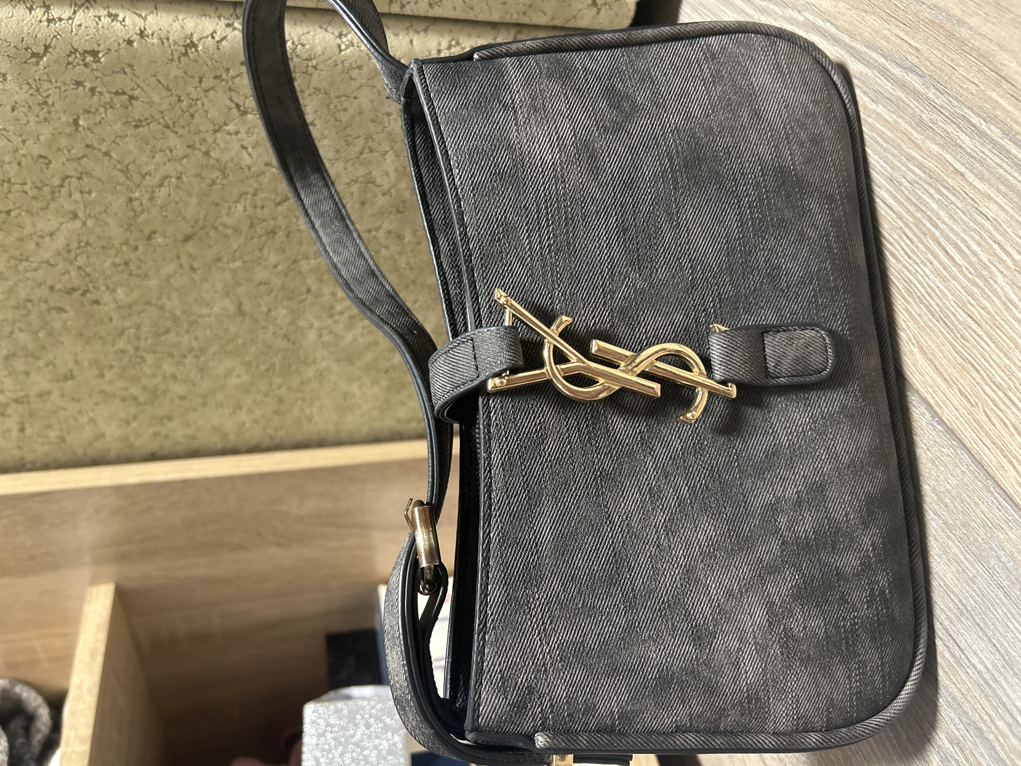 A dark grey denim shoulder bag with a gold Yves Saint Laurent logo is displayed on a wooden surface. The bag has a single shoulder strap and a flap closure. The background consists of a wooden shelf and a textured wall. The scene is indoors, and there are no people or animals present. The image was taken on a flat lay from a slightly elevated angle. The lighting is natural, coming from an unseen source. The overall environment appears to be a domestic setting, possibly a bedroom or closet. There is no indication of the city of Ivanivka, Ukraine in the image.