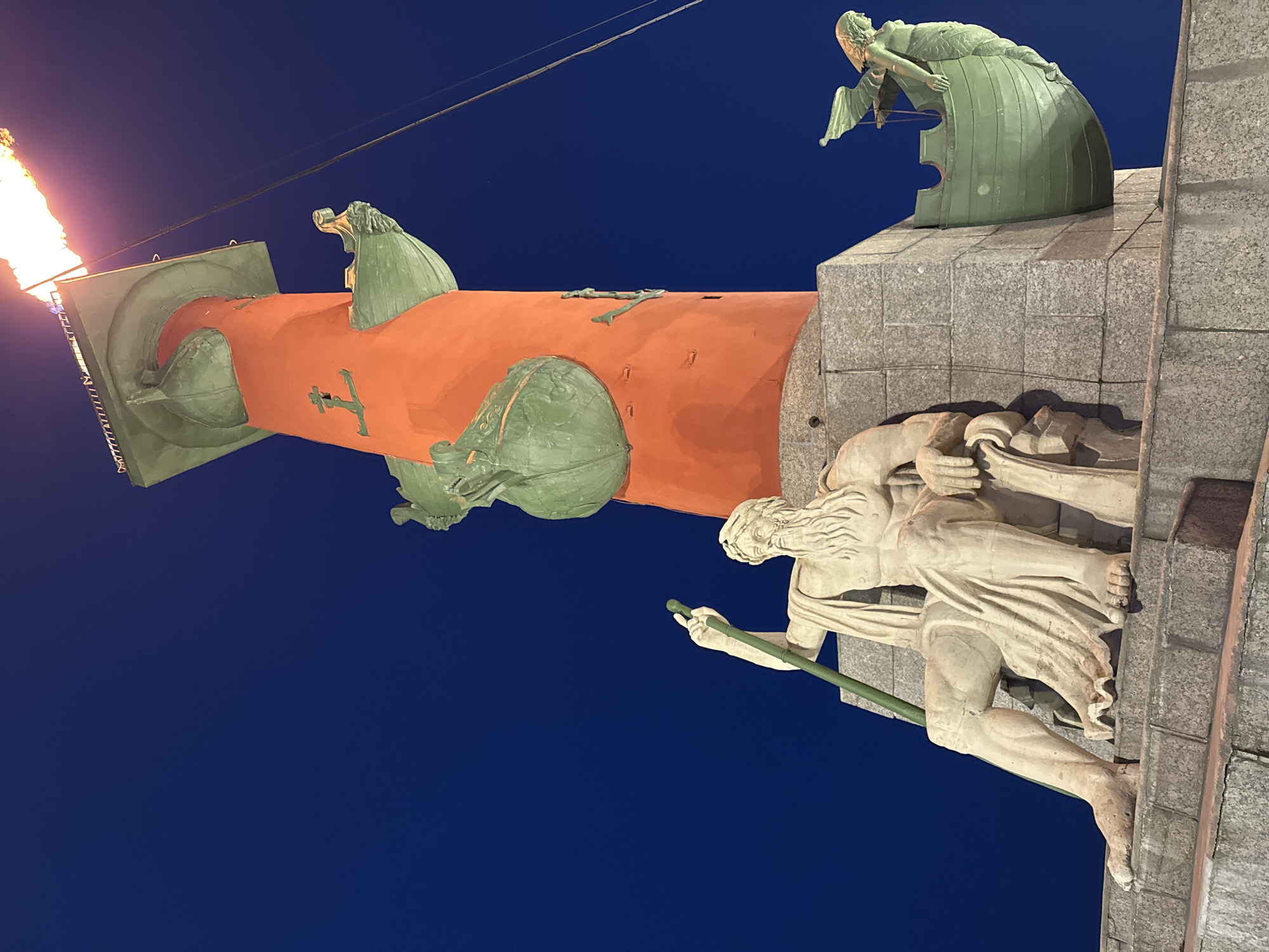 The image captures a nighttime view of the Rostral Columns in Saint Petersburg, Russia. The columns are adorned with green nautical figureheads. One column has a flame burning at the top. A stone sculpture of a male figure holding a staff is near the column base. The sky is a deep blue.