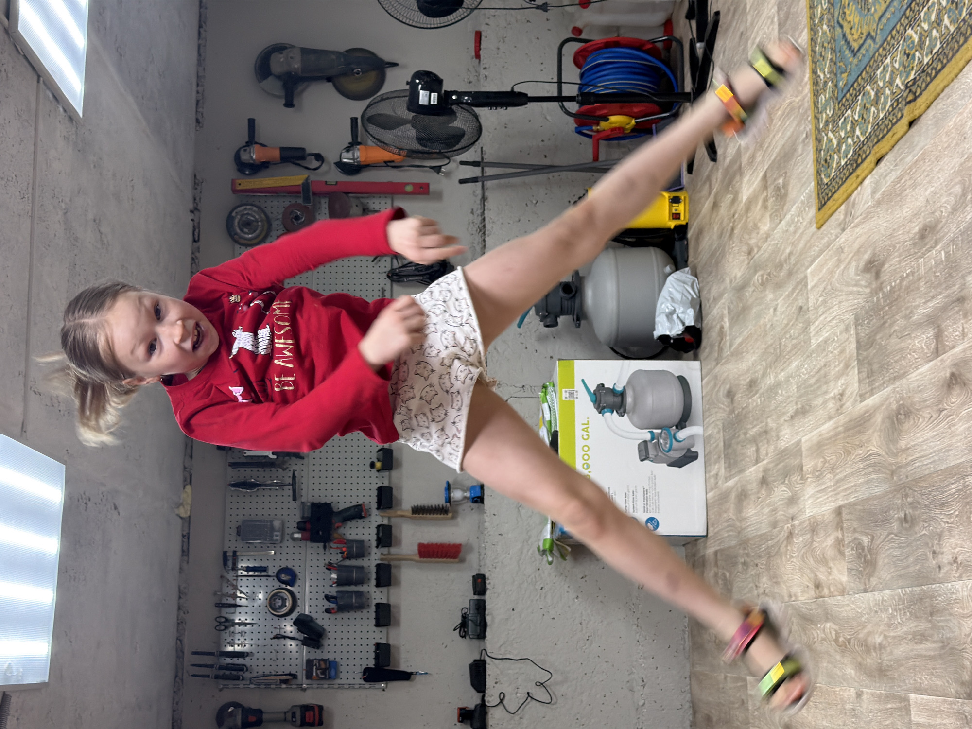 A young girl in a red long-sleeved shirt and patterned shorts is performing a high kick in what appears to be a basement or workshop. She wears colorful sandals on her feet. Various tools are hung on the wall behind her, including drills, grinders, and other equipment. A fan and a hose reel are also visible. A gray water tank, and its box is located near the wall, as well as a rug in the corner. The location is a workshop in Dubovaya, Russia.
