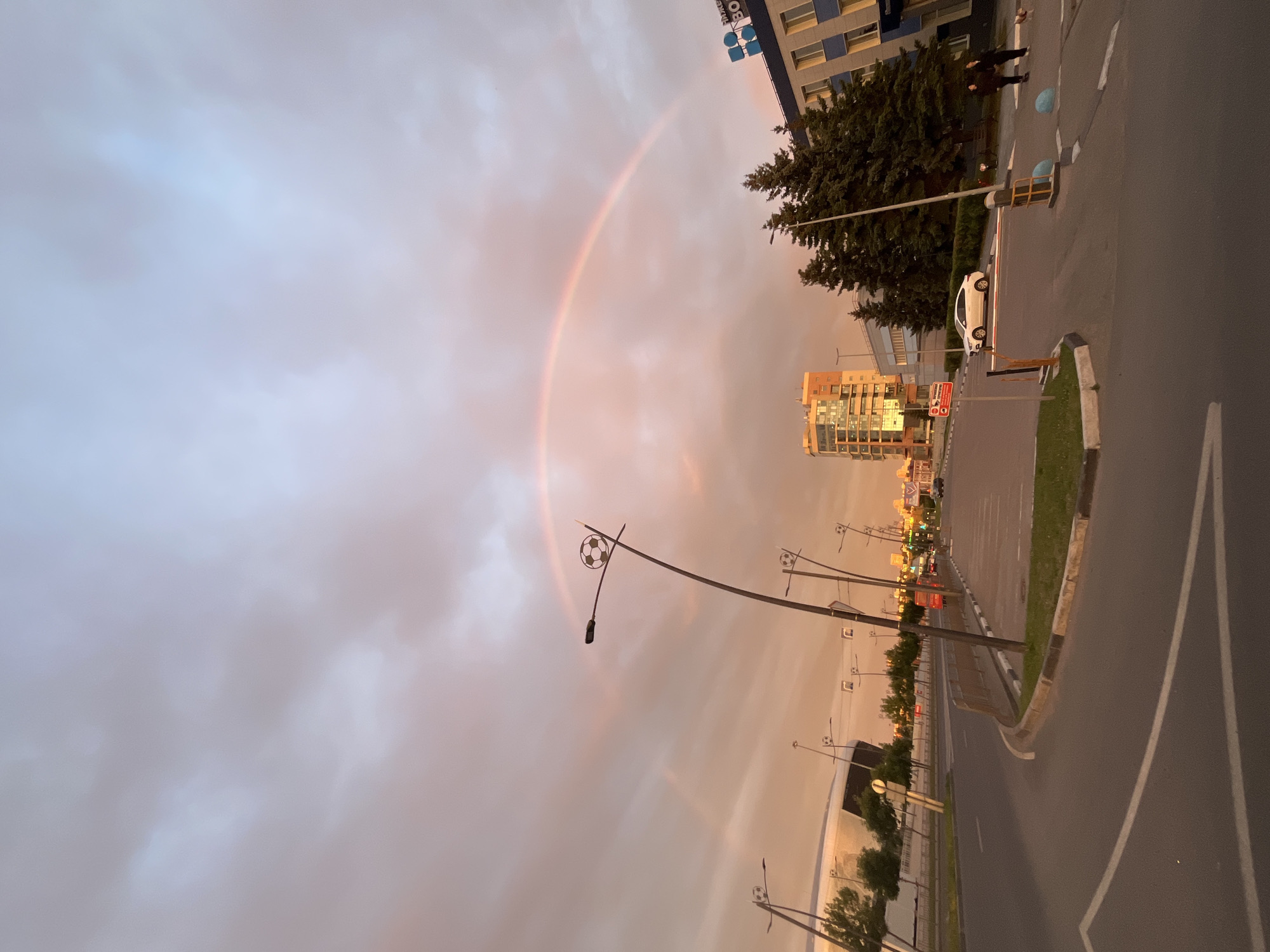 The image depicts a street scene at dusk in Nizhny Novgorod, Russia. A prominent rainbow arcs across the overcast sky, casting a warm, peachy glow. The sky is filled with soft, diffused clouds, suggesting recent or ongoing rain.
The scene includes a road with a white car parked on the right side. A few streetlights are visible, some adorned with decorative soccer ball motifs. Beyond the road, there are trees, a building with a blue facade and white windows, and another taller building with a more modern glass exterior. Two people are standing on the sidewalk near the blue-facade building, appearing to observe the rainbow.
The overall atmosphere is tranquil and somewhat magical due to the presence of the rainbow and the soft evening light. The time of day is clearly sunset or twilight, indicated by the warm tones in the sky and the dimming light. The presence of a street and buildings suggests an urban or suburban setting. No clear text is legible in the image, except for a sign on a pole that is too small to read.