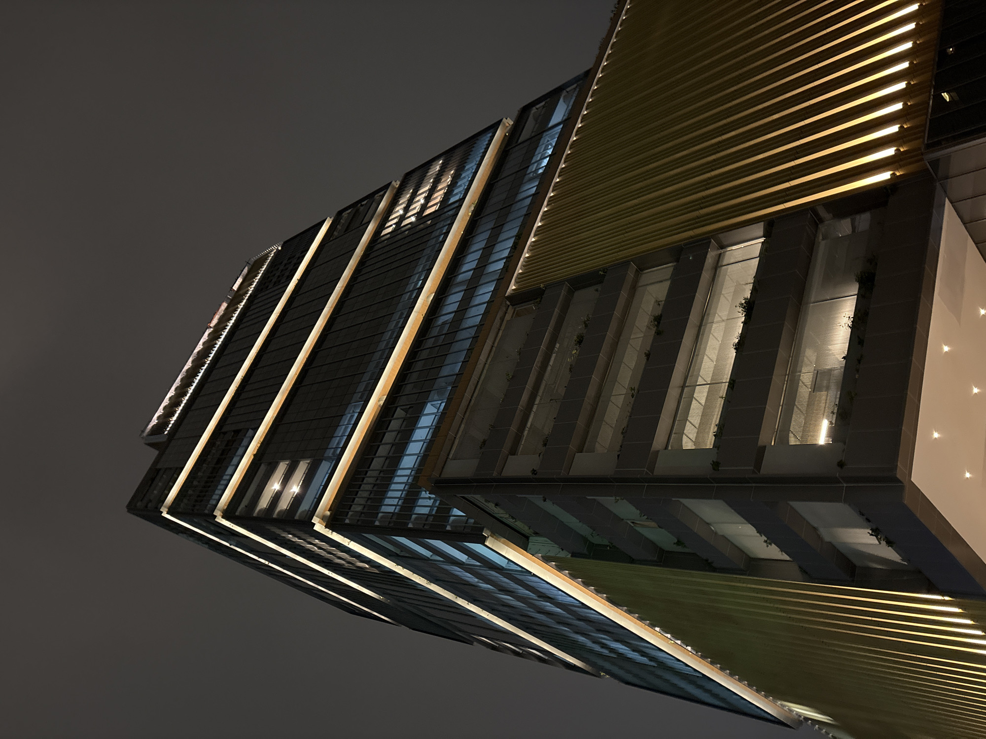 A low-angle, full shot shows the exterior of a modern skyscraper at night, possibly in the city of São Paulo, Brazil. The building has a dark, reflective glass facade with horizontal bands of warm, golden light running along its length, highlighting its verticality. Part of the building features a ribbed, gold-colored cladding, also illuminated from below by vertical lights. Several floors are visible, with some windows showing interior lights and the silhouettes of plants. The sky above is dark and overcast, indicating nighttime. No people are visible.