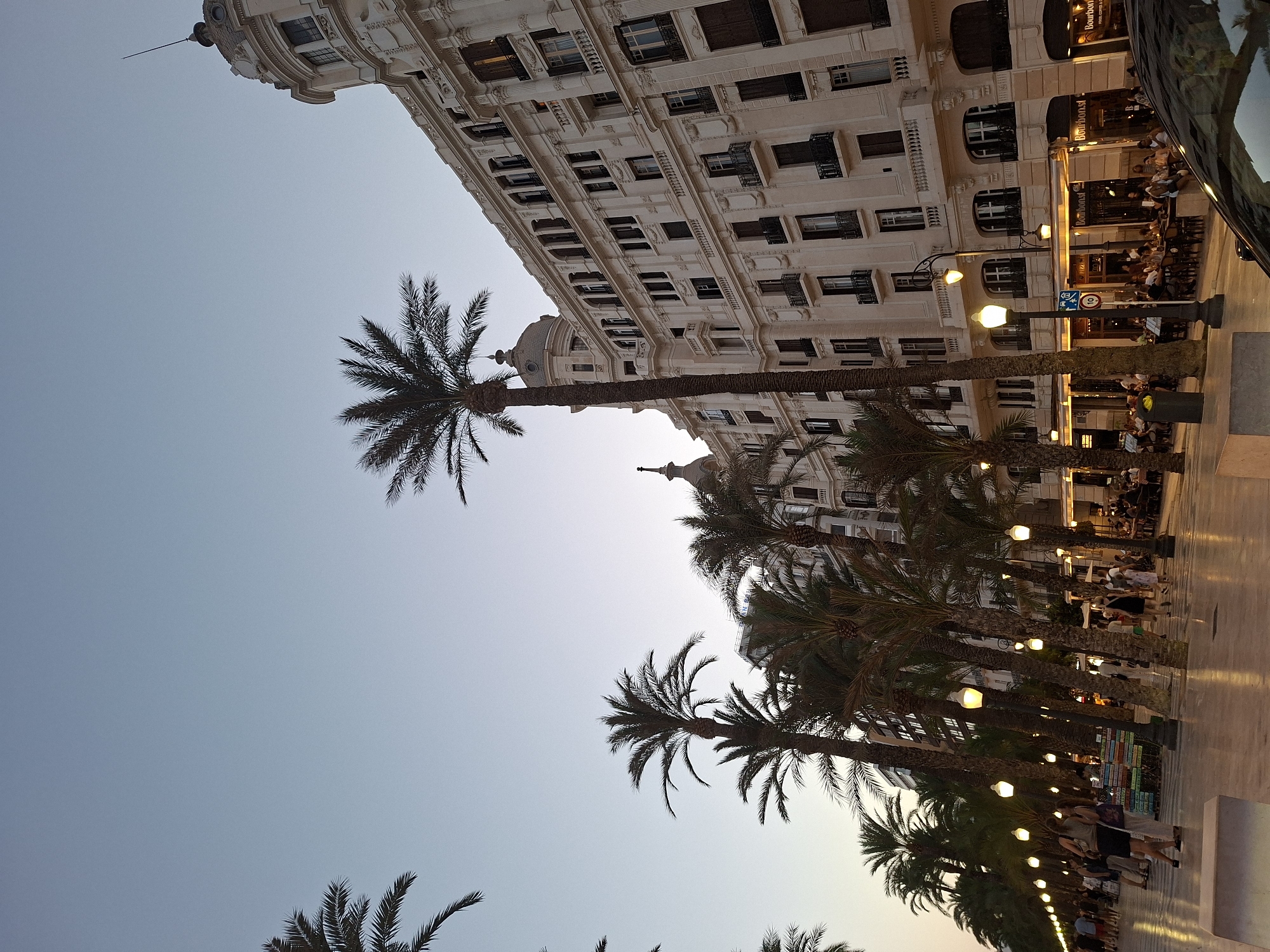 The image shows the Explanada de España, a promenade in Alicante, Spain. It features a wide pedestrian walkway lined with palm trees on the left and a large, ornate, cream-colored building on the right. The building has multiple stories with many windows and balconies, and architectural details characteristic of early 20th-century design, including domes and spires. In the foreground, people are seen walking along the promenade and sitting at tables outside cafes and restaurants on the ground floor of the building. The lighting suggests it is late afternoon or early evening, with streetlights illuminating the walkway. The sky is clear and a pale blue.