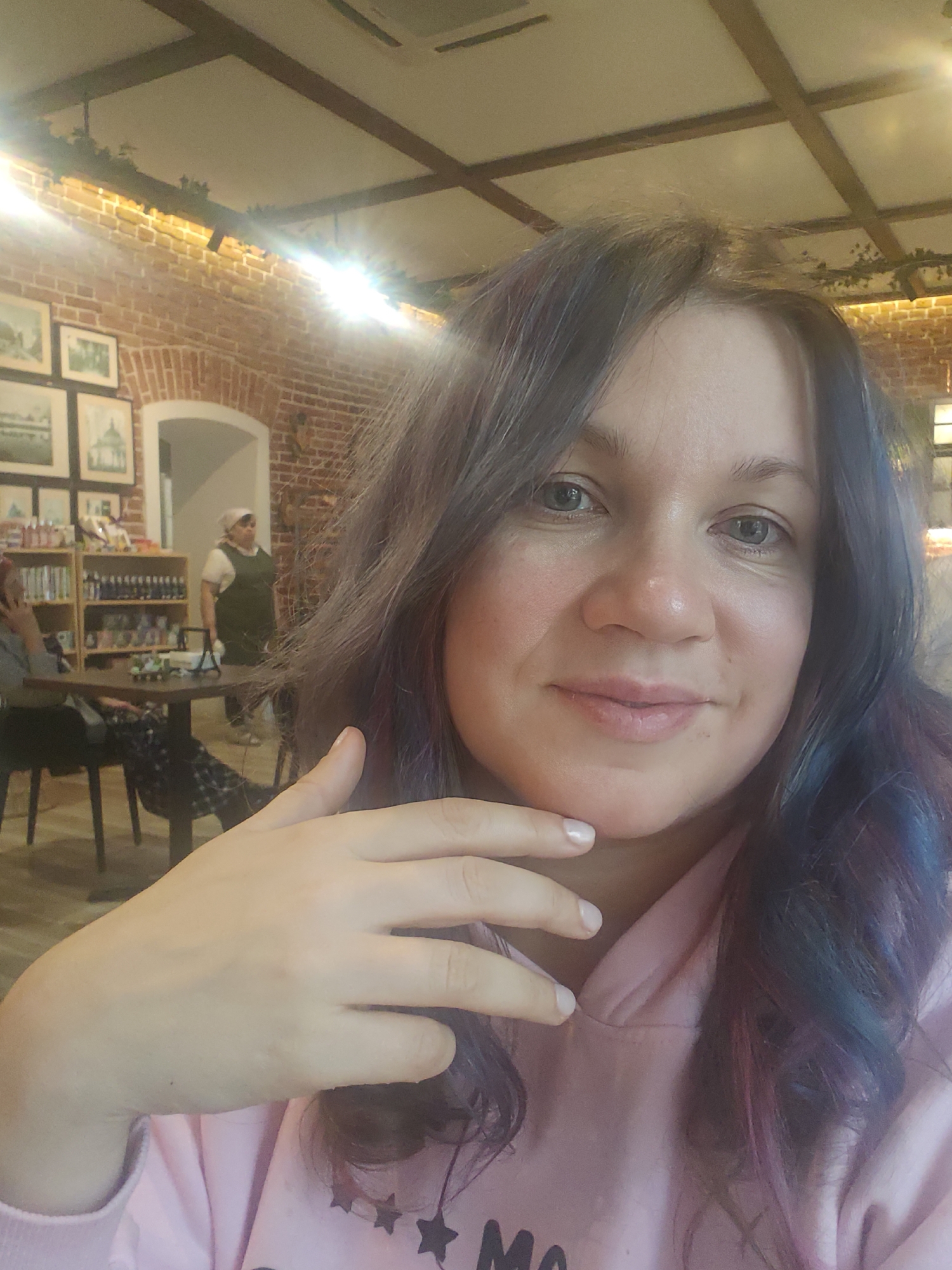 A medium shot captures a woman in what appears to be a café. Her gray hair is styled with waves, and there are visible hints of blue and purple. She is wearing a light pink hooded sweatshirt. Her right hand is raised to her chin, with her fingers close together.
The background reveals an interior with brick walls, wooden beams in the ceiling adorned with greenery, and framed pictures. Other patrons and staff can be seen in the background, along with shelves of various products. The location is Куликово, Russia.