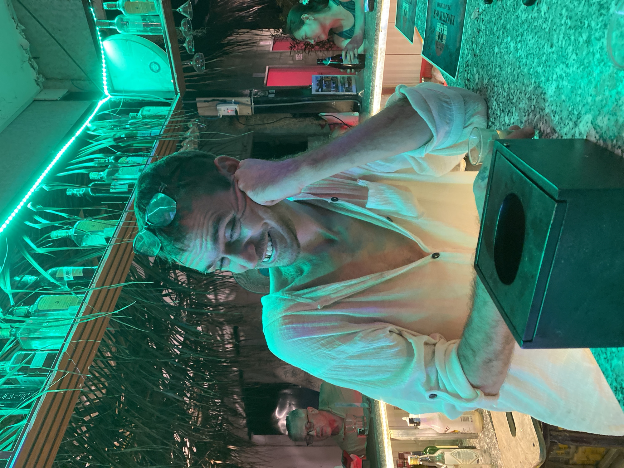 This image depicts a vibrant bar scene in Mũi Né, Vietnam, illuminated predominantly by green light.
In the foreground, a Caucasian male, estimated to be in his 30s-40s, is seated at a bar counter. He has short, dark hair, light stubble, and sunglasses resting on top of his head. He is wearing a light-colored, possibly off-white or cream, long-sleeved collared shirt, unbuttoned to reveal his chest, with sleeves rolled up to the forearms. He is smiling broadly, with his head tilted to his right (viewer's left), and his left hand supports his face at the cheek/ear. His left forearm rests on the bar counter. To his right, on the counter, is a small clear glass containing a light-colored liquid. Directly in front of him is a dark, rectangular object, possibly a tissue dispenser or menu holder. The bar counter itself is a mottled green color, likely granite or a similar stone, reflecting the green light.
The immediate background reveals more of the bar setup. Above the main subject's head is a structure composed of wooden slats and dense, dried palm fronds or similar foliage, creating a thatched-roof effect. A string of green LED lights runs along the top edge of this structure, casting the pervasive green hue throughout the scene. Various liquor bottles, including one labeled "MARIBU" (Malibu), are visible on shelves above the foliage. Inverted stemware hangs from racks further to the right.
To the far left in the background, another Caucasian male, appearing older (possibly 50s-60s) with glasses and a light-colored shirt, is seated at the bar, facing towards the right. A glass is in front of him on the counter. The wall behind him appears to be made of stone or textured plaster.
To the far right in the background, an Asian female, appearing younger (possibly 20s-30s) with dark hair tied back, is also seated at the bar. She wears a dark tank top and holds a dark bottle, possibly a beer bottle, in her right hand. She is looking towards the left. Behind her, a red door or entryway is visible, along with white rectangular signage on the counter. Between the main subject and the female in the background, a white electrical box with switches is mounted on a post.
The overall environment is that of a casual, tropical-themed bar, vibrant with the distinctive green lighting.