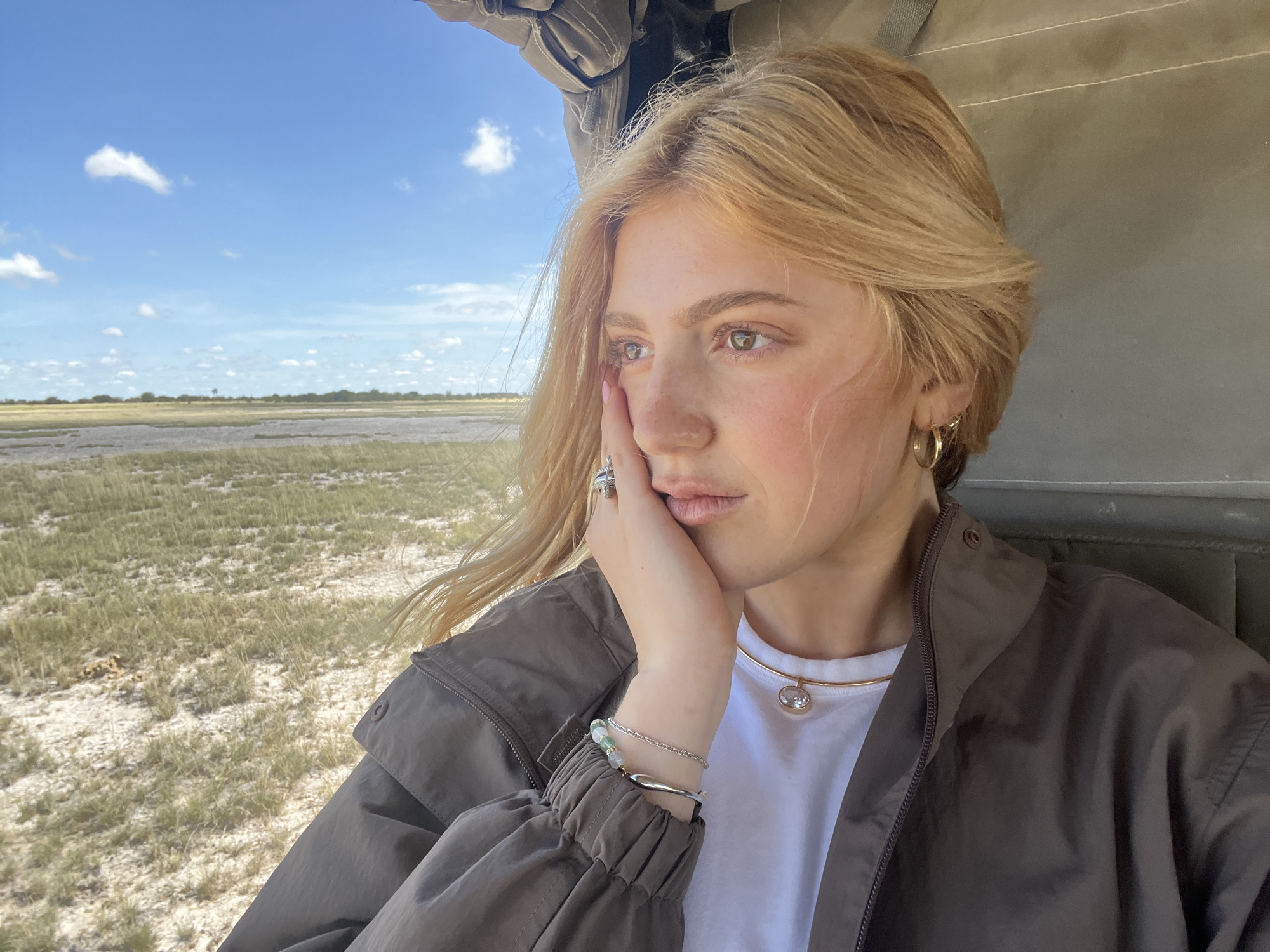 A woman with blonde hair, wearing a brown jacket over a white shirt, is looking out of a window. She has jewelry on her fingers, wrist, and neck. The background outside the window shows a flat landscape with vegetation and a blue sky with clouds. It appears to be the Okavango Delta, near Gucta, Botswana.