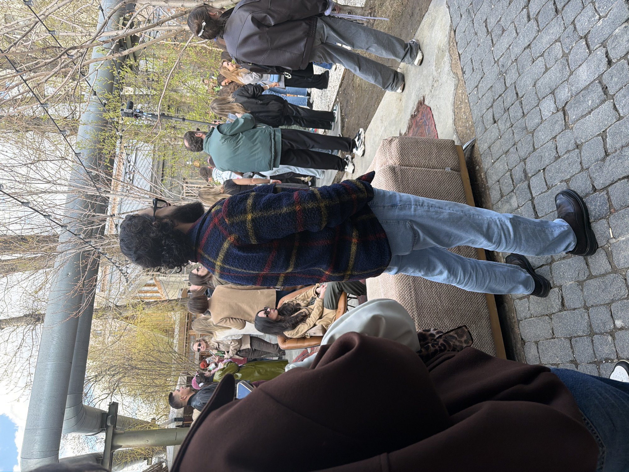 The photograph depicts an outdoor scene with a gathering of people. The setting appears to be an urban area with exposed pipes and bare trees. There's a diverse group of individuals present, some standing in a line and others seated or standing around in a more casual arrangement. Clothing styles vary, including jackets, sweaters, jeans, and dresses. A cobblestone pathway is visible in the foreground, along with sections of outdoor seating, perhaps a patio or public area. The location could be in Moscow, Russia, given the architectural details and urban setting.