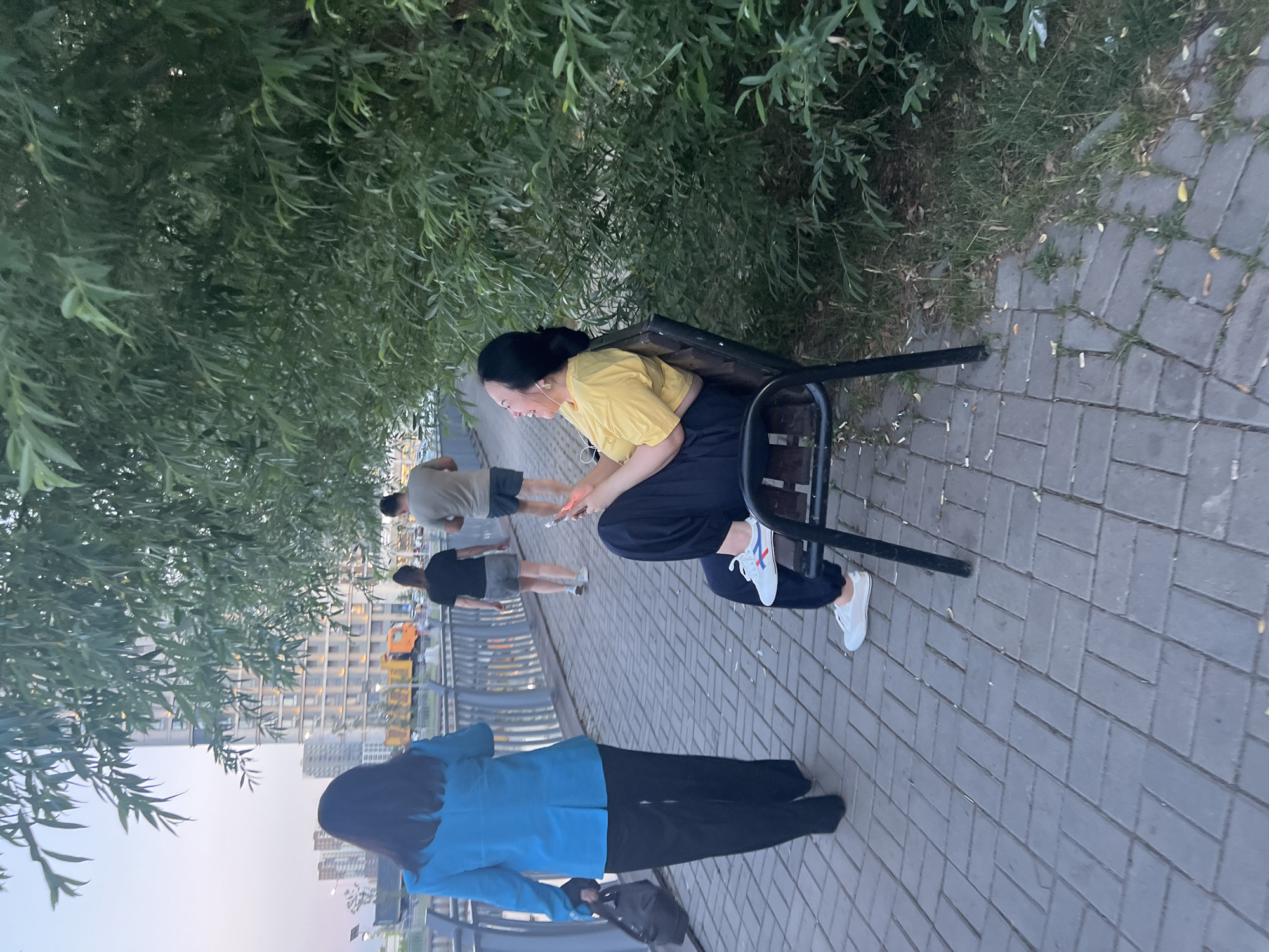 The image depicts a paved walkway alongside a body of water, likely a river or canal, in Astana, Kazakhstan. On the walkway, a woman wearing a blue blazer and black pants walks away from the camera, carrying a black bag. Further down the path, two individuals, a woman and a man, are walking away from the camera, with the man in a grey t-shirt and shorts, and the woman in a black t-shirt and shorts. To the right of the walkway, a woman sits on a bench, wearing a yellow top and dark pants, looking to her left and holding a smartphone. She has earbuds in her ears and appears to be smiling. The area is lined with lush green trees and foliage. In the background, modern buildings and city structures are visible. The ground is paved with rectangular grey bricks laid in a herringbone pattern. The lighting suggests it is either early morning or late evening.