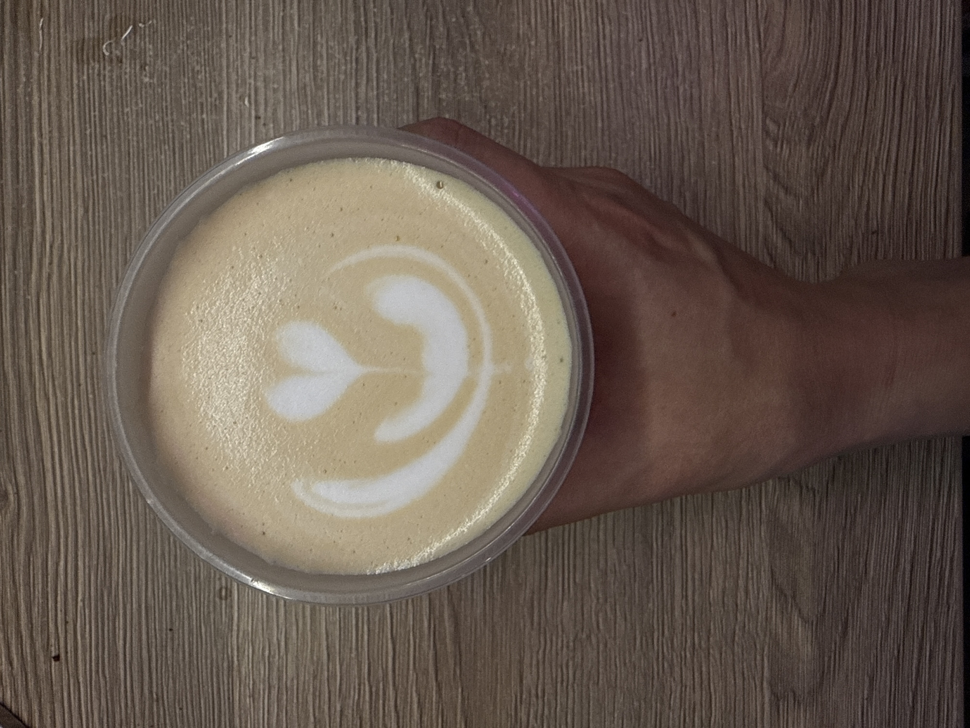 A clear plastic cup of coffee with latte art in the shape of a heart and a petal design is held by a hand against a wooden surface. The coffee has a creamy texture with a foamy top. The wooden surface appears to be a table with a visible wood grain pattern. The image was taken in Veshki, Russia.
