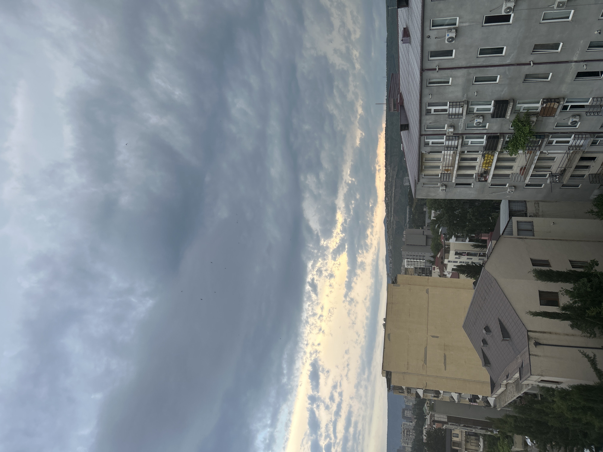 This media file captures a view of buildings and a cloudy sky. The perspective suggests the photo was taken from a height, looking down and across a cityscape.
The main subjects are apartment buildings with multiple floors, characterized by numerous windows, balconies, and visible air conditioning units. One building is a light gray, while another is a pale yellow. There are glimpses of other buildings and trees in the background, suggesting a densely populated urban or suburban area. The sky is filled with a mixture of dark and lighter gray clouds, with some areas illuminated by sunlight peeking through, creating a dramatic and somewhat overcast atmosphere. A few birds are visible in the sky, appearing as small dark specks.
The time of day appears to be either early morning or late afternoon, given the soft, diffused light and the color of the sky. The weather seems to be transitioning, with clouds suggesting potential rain or simply a cloudy day. The location is not explicitly identifiable, but the architecture of the buildings and the presence of greenery hint at a temperate climate. There is no discernible activity or event taking place, other than the passive presence of the buildings and the natural occurrence of the weather. The overall mood evoked by the image is one of quiet observation of an urban environment under a dynamic sky. There is no visible text within the image.