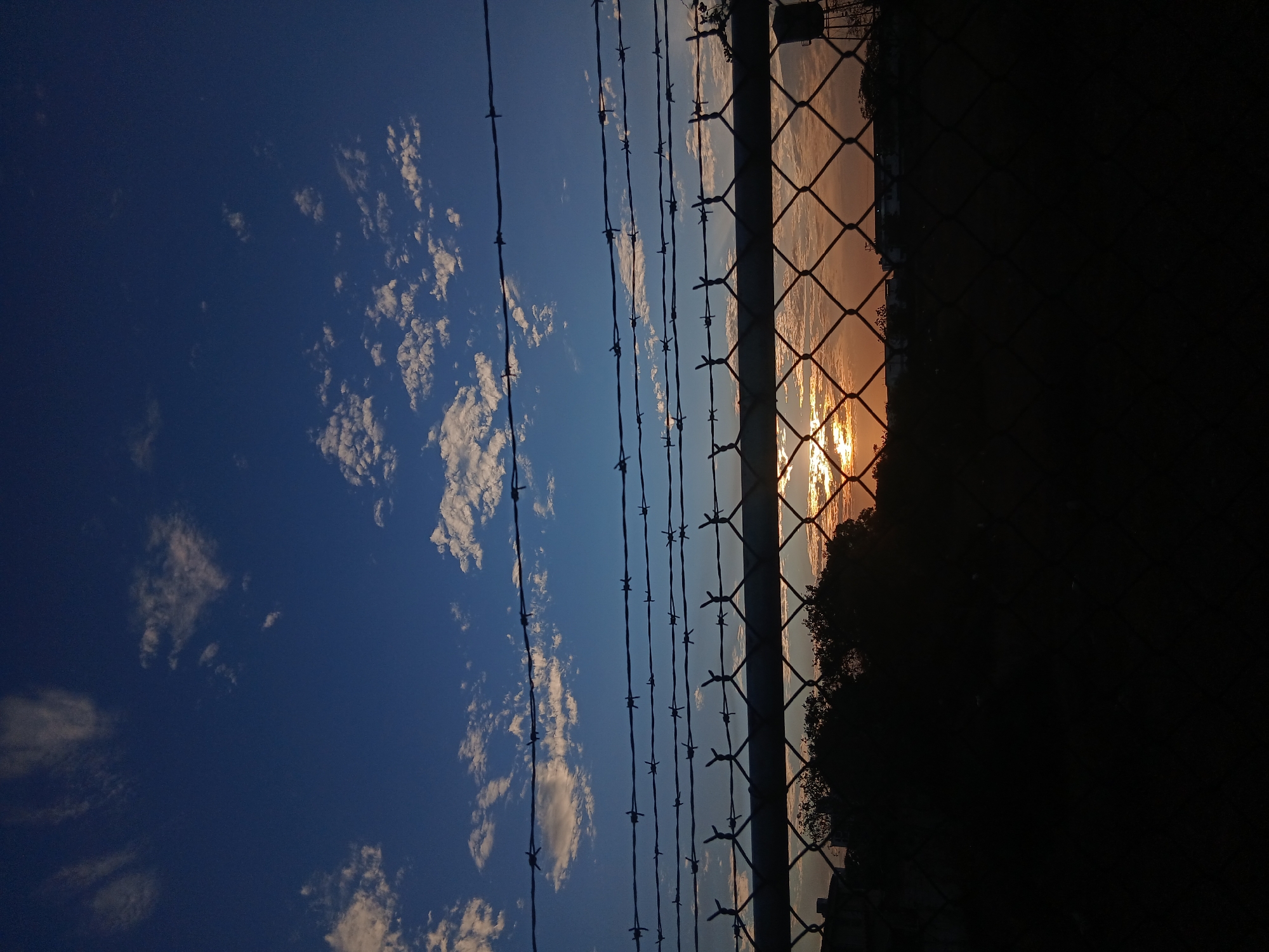 The photograph depicts a scene at sunset in Juan Griego, Venezuela. A chain-link fence and strands of barbed wire are in the foreground, partially obscuring the view. The sky transitions from deep blue with scattered white clouds on the left to orange and golden hues on the right, where the sun is setting. Silhouetted trees are visible behind the fence, and darkness encroaches on the right side of the image. There are no people visible in the photograph.