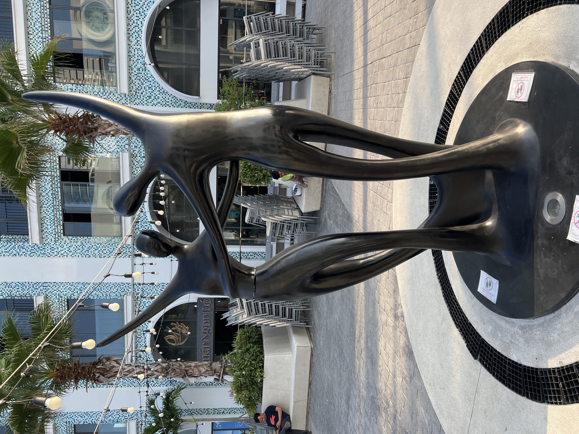 A black, abstract sculpture of two intertwined figures dancing is positioned in a paved outdoor area in Nha Trang, Vietnam. The sculpture is centrally located on a circular platform with a black grate surrounding it. In the background, a modern building with blue and white tiled facade and arched windows is visible, along with palm trees. Several stacks of empty chairs are arranged in front of the building, suggesting an outdoor dining or seating area. A man in a blue shirt is sitting on a concrete bench to the left, looking at his phone. Another person wearing a yellow vest is visible further back, near the chairs. String lights are hung overhead, illuminating the scene. Two small signs with prohibition symbols are attached to the base of the sculpture.