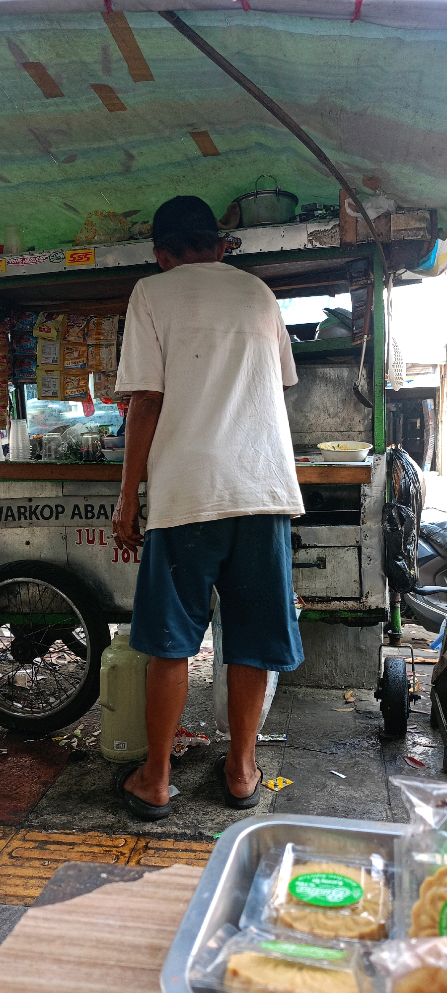 An Indonesian man stands with his back to the camera, wearing a white t-shirt and blue shorts, at a food cart in Bandung City, Indonesia. The cart has a green metal frame and is covered with a green tarp, patched with brown tape. Several packets of snacks are displayed on a shelf inside the cart. A large green water jug sits next to the cart. In the foreground, a metal tray contains individually wrapped pastries, some with green labels. The ground appears to be paved with concrete and there is some debris. A motorcycle is partially visible to the right of the frame. The overall scene suggests a typical street-side food vendor operation.
