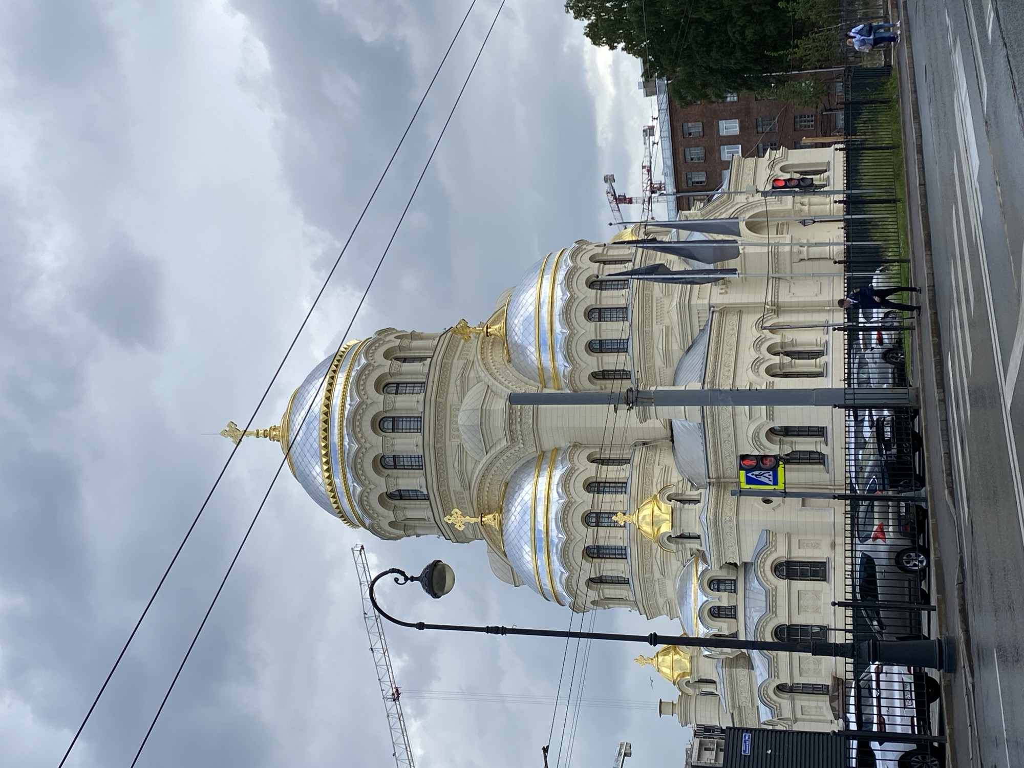 The image shows the Naval Cathedral of Saint Nicholas in Kronstadt, a city within Saint Petersburg, Russia, on an overcast day. The large, cream-colored cathedral features multiple domes, with the central and smaller ones topped with gold crosses and silver plating. The cathedral has numerous arched windows and decorative elements.
In the foreground, a wet street with a pedestrian crossing is visible. Several cars are parked along the sidewalk behind a black metal fence. A black lamppost with an ornate design stands on the left side of the image. A pedestrian traffic light shows a red signal for pedestrians.
A man in a dark uniform is walking on the sidewalk, and two women are walking further down the sidewalk. In the background, several construction cranes are visible, suggesting ongoing development or renovation in the vicinity. Trees are present on the right side of the image, and an apartment building with brick facade is situated behind the cathedral. Electrical wires are strung across the sky.