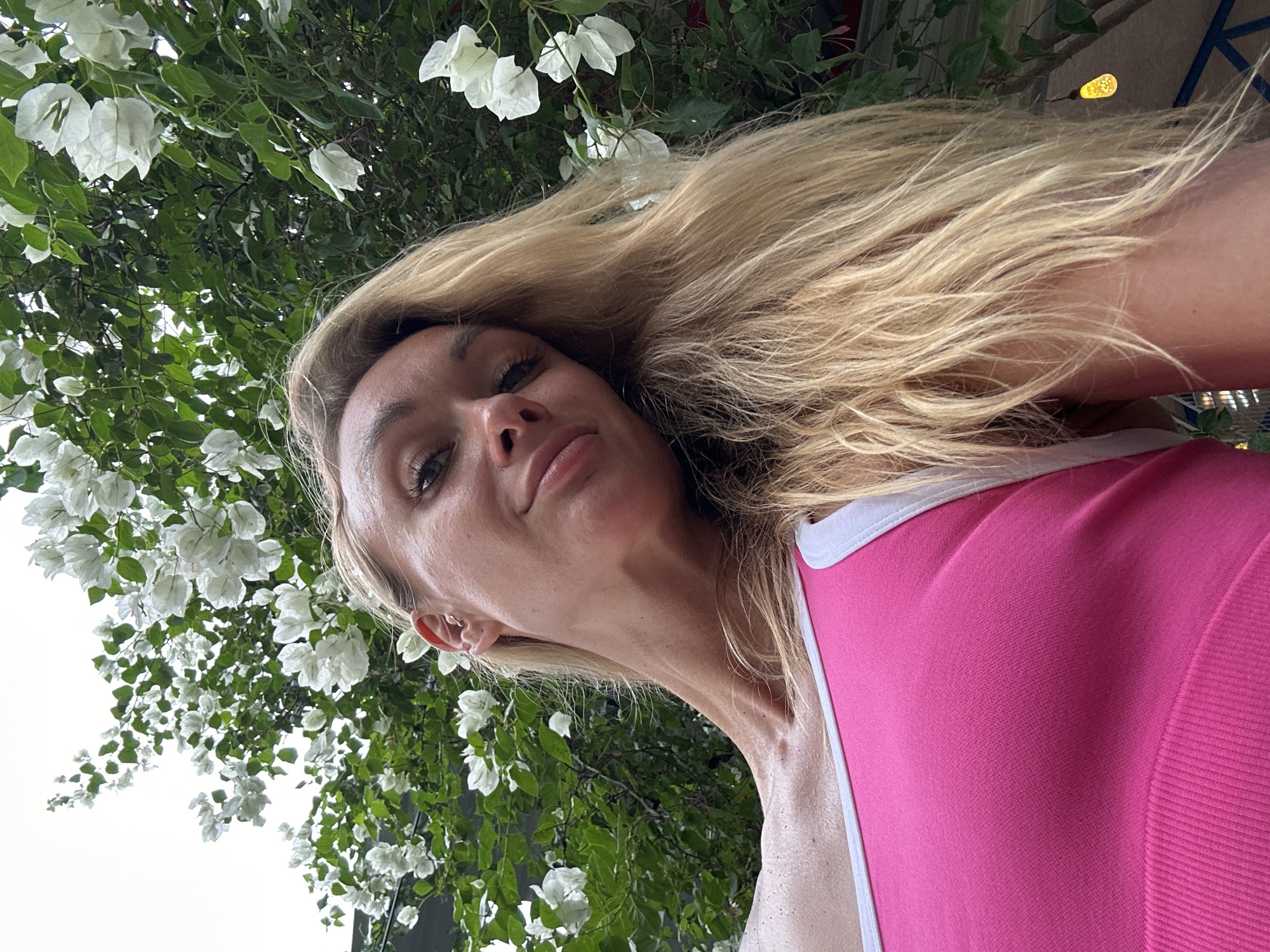 A young woman with blonde hair is photographed from a low angle, looking upwards towards the camera. She is wearing a pink tank top with white trim. Behind her, a tree with abundant white flowers and green leaves is visible against a bright sky. The image appears to be taken outdoors in a location that suggests lush vegetation, possibly in the city of Phan Sơn, Vietnam, although this cannot be definitively confirmed from the visual information alone. There are no other individuals or significant objects in the frame besides the woman, the flowering tree, and the sky. The woman's expression is relaxed and she is looking directly at the camera.