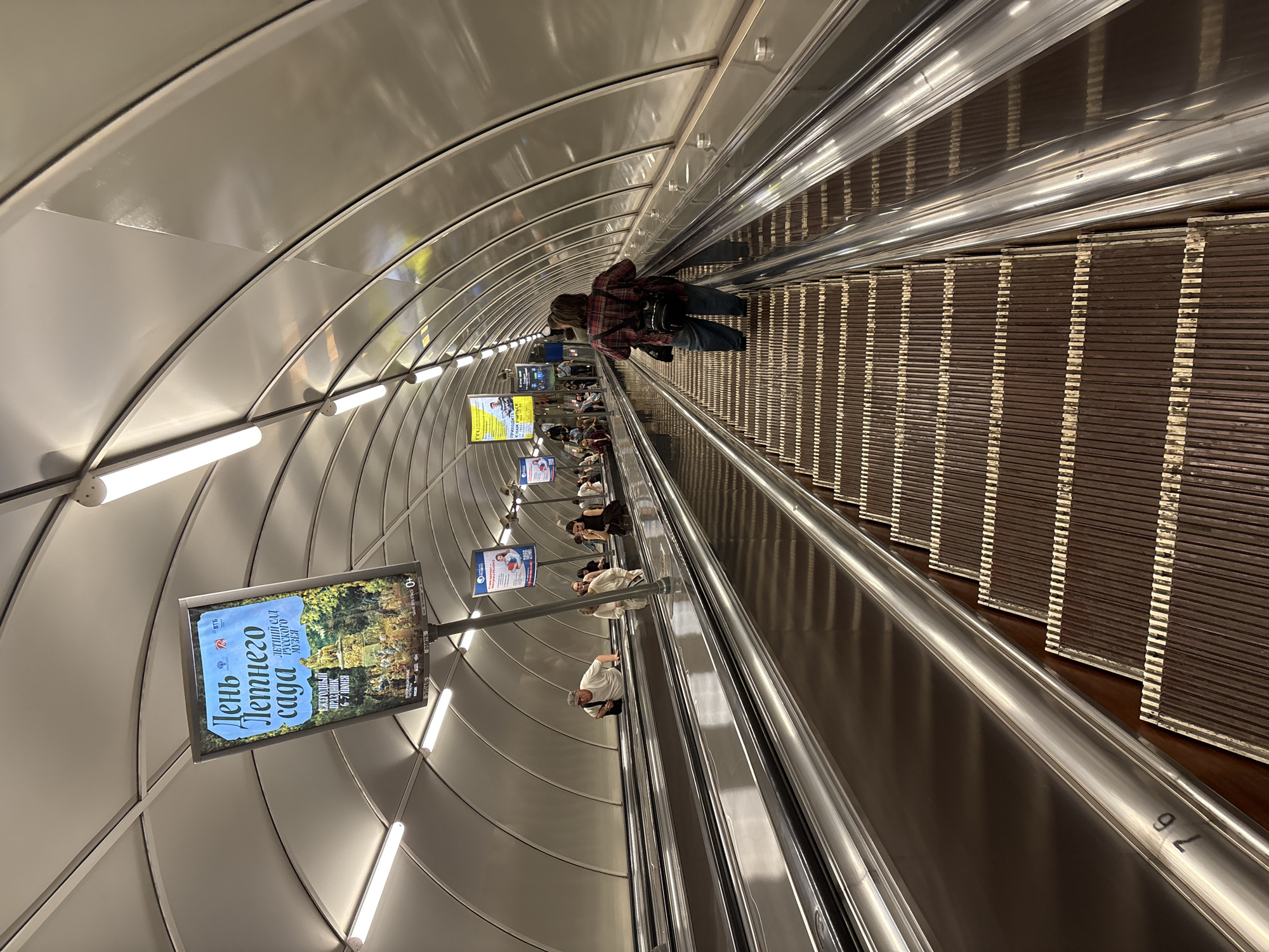 The image shows an escalator going down in what appears to be a subway station. The escalator is filled with people going down. On the side of the escalator are advertisements, one of which is for the "Day of the Summer Garden" on June 6-7. The ceiling of the station is curved and lined with lights. A number "76" is visible on the right.