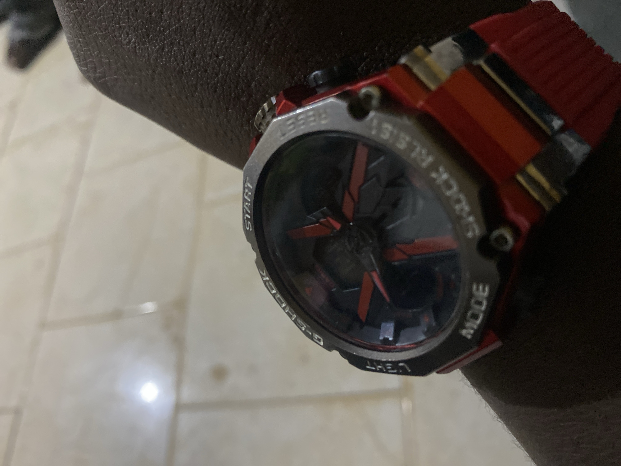 A close-up, slightly angled shot captures a person's left arm and hand, adorned with a distinctive red and silver G-Shock style watch. The watch features a robust silver bezel with visible text engravings: "START" on the left, "RESET" at the top, "SHOCK RESIST" on the upper right, "MODE" on the lower right, "LIGHT" at the bottom, and "G-SHOCK" on the lower left. The watch face is dark, with prominent red accents forming a geometric, almost propeller-like design in the center. While the watch appears to be an analog-digital model, the digital display is not clearly visible or illuminated. The watch strap is a vibrant red, contrasting with the silver bezel.
The arm, exhibiting a dark skin tone, is positioned with the watch prominent. The background reveals an indoor setting with light-colored, possibly beige or off-white, tiled flooring. A bright light source creates a noticeable glare on the reflective surface of the tiles, suggesting artificial illumination, which could imply evening or night, or simply an indoor environment in Shen, Nigeria with limited natural light. The focus is primarily on the watch and the arm, with the background slightly out of focus. No specific activity is captured beyond the person wearing the watch.