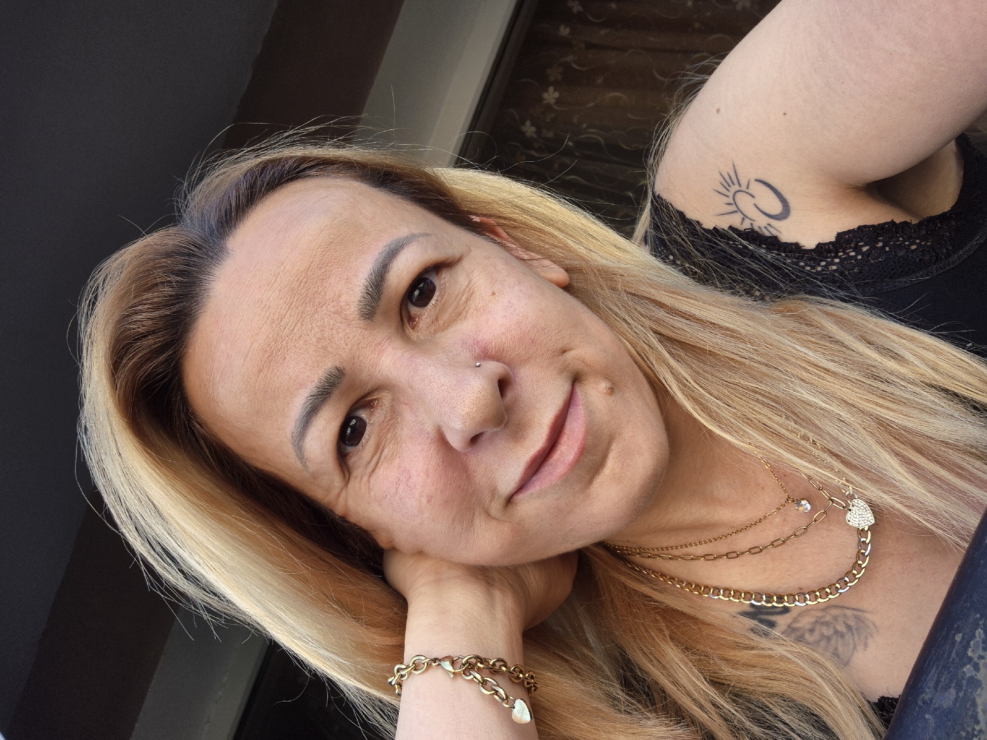 A close-up, medium shot captures a fair-skinned individual from the shoulders up, facing the camera with a slight smile. They have blonde, shoulder-length hair with subtle blonde highlights. Their left arm is bent, with the hand resting behind their neck, adorned with a gold bracelet featuring two heart-shaped charms. They wear a black top with lace detailing visible on the shoulder and chest. Two gold necklaces are layered around their neck, one thicker chain and a thinner one with a small, clear pendant. A tattoo of a crescent moon with sun rays is visible on the upper right shoulder. A small nose ring is present on the left side of their nose. The background is blurred, showing what appears to be an outdoor setting with a dark, textured surface above and a patterned curtain or screen behind them. The lighting suggests it is daytime.