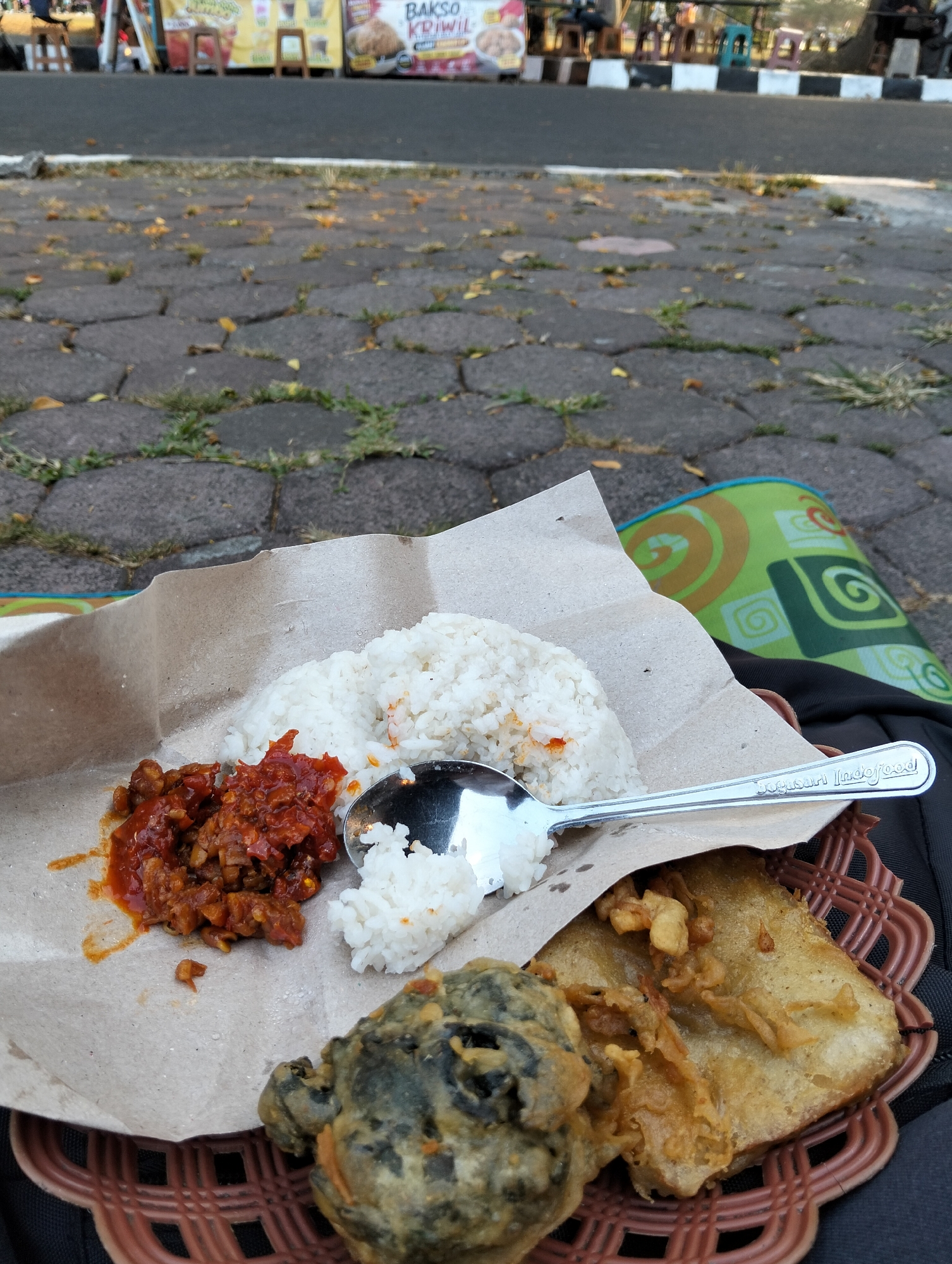 The image depicts a close-up view of a meal served on brown paper, placed within a woven basket. The meal consists of white rice, a portion of red chili paste or sauce with meat, and two pieces of fried food. One fried item appears to be a large piece of tempeh, while the other is a dark, irregularly shaped item, possibly a piece of fried mushroom or other vegetable. A metal spoon, engraved with "Jugusan Indofood", rests on the rice.
The basket is situated on a dark surface, possibly a backpack, which is in turn placed on a cobblestone ground interspersed with patches of grass and scattered leaves. In the background, out of focus, is a street scene with a sidewalk and a row of shops or food stalls. Banners with text are visible, one of which clearly displays "BAKSO KRIWIL". The lighting suggests it is daytime, with shadows present. The scene is likely set outdoors in an urban environment in Magelang, Indonesia, given the visual cues of street food stalls and the specific text on one of the banners.