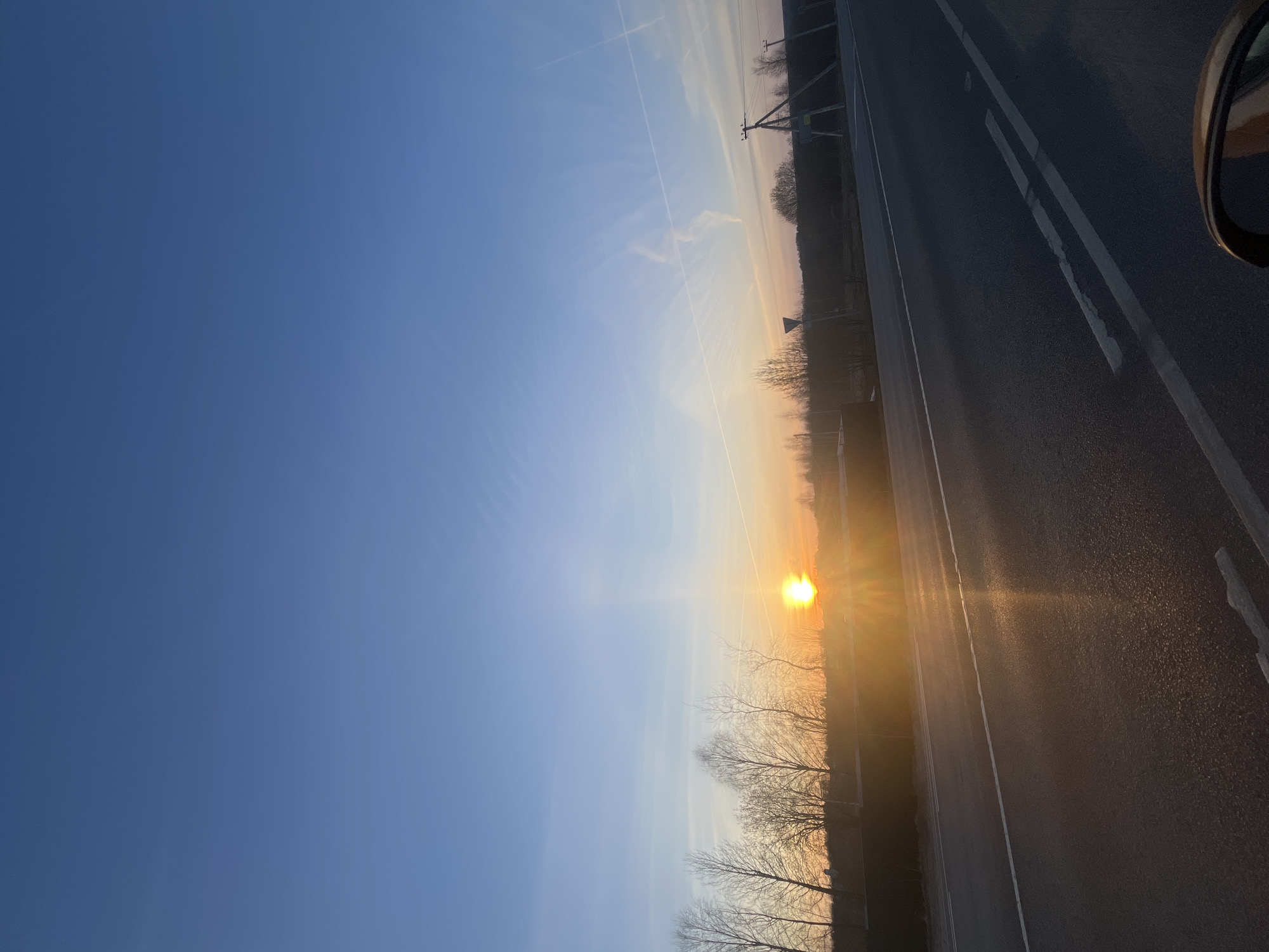 The image depicts a sunset on a road, likely taken from a moving vehicle. The sun is low on the horizon, casting a warm glow across the asphalt and illuminating the bare branches of trees lining the roadside. A clear blue sky transitions to orange and yellow hues near the horizon. A portion of a car's side mirror is visible in the upper right corner of the image. The photograph was taken in Zaovrazh'e, Russia. There are no people visible in the photograph.