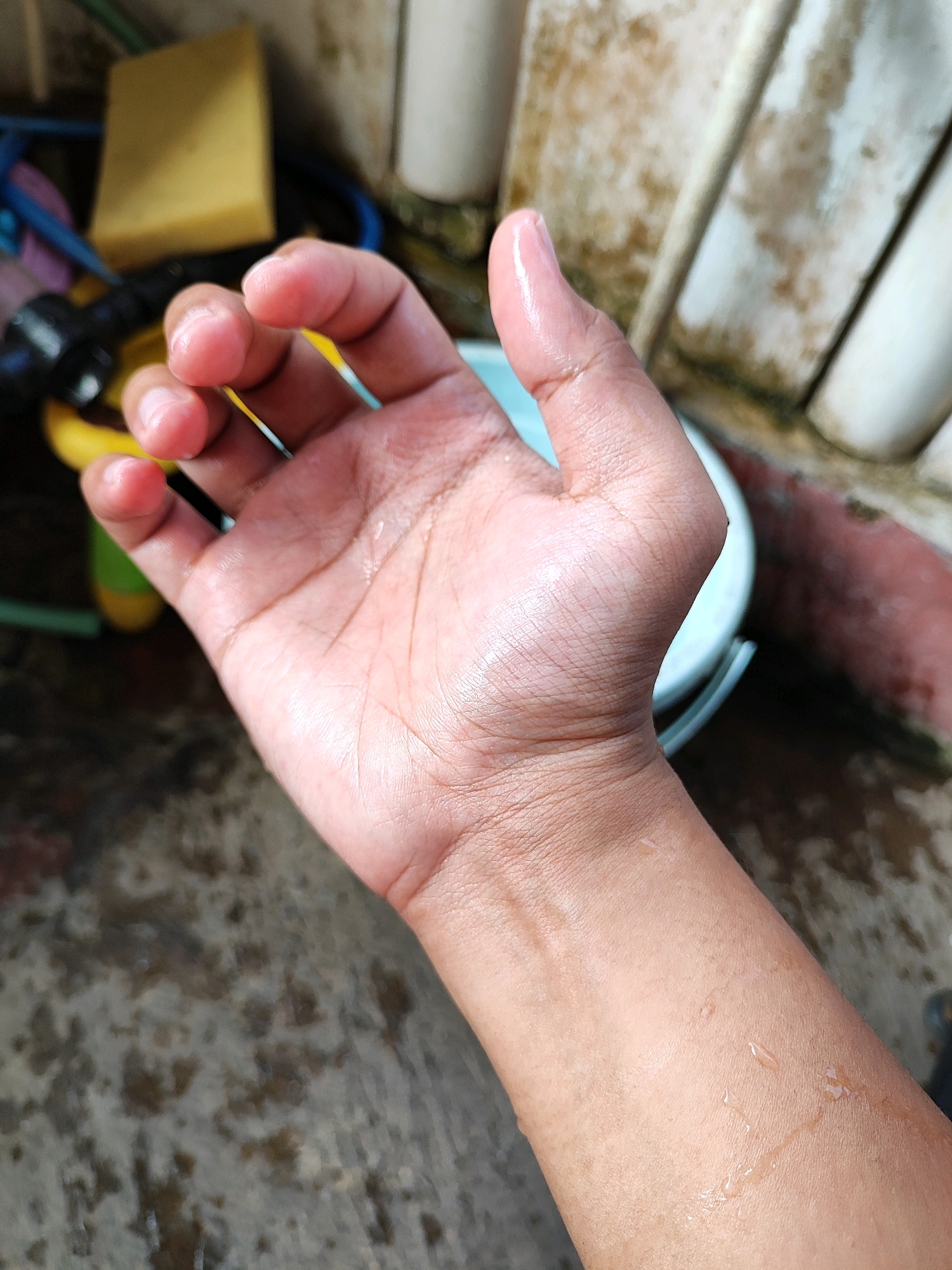 A close-up, overhead shot captures a left forearm and hand with visible water droplets on the skin, suggesting it is wet. The skin tone is light brown, with fine wrinkles and lines apparent. The fingers are slightly curled inward, and the thumb is resting on the palm. In the background, out of focus, are various objects including a yellow sponge, blue and black hoses, and a light blue bucket. The ground is a mottled, wet surface that appears to be concrete or paving stones. The overall scene suggests an outdoor setting, potentially in a yard or utility area. The specific location within Bondowoso, Indonesia, cannot be determined from the visual information provided.