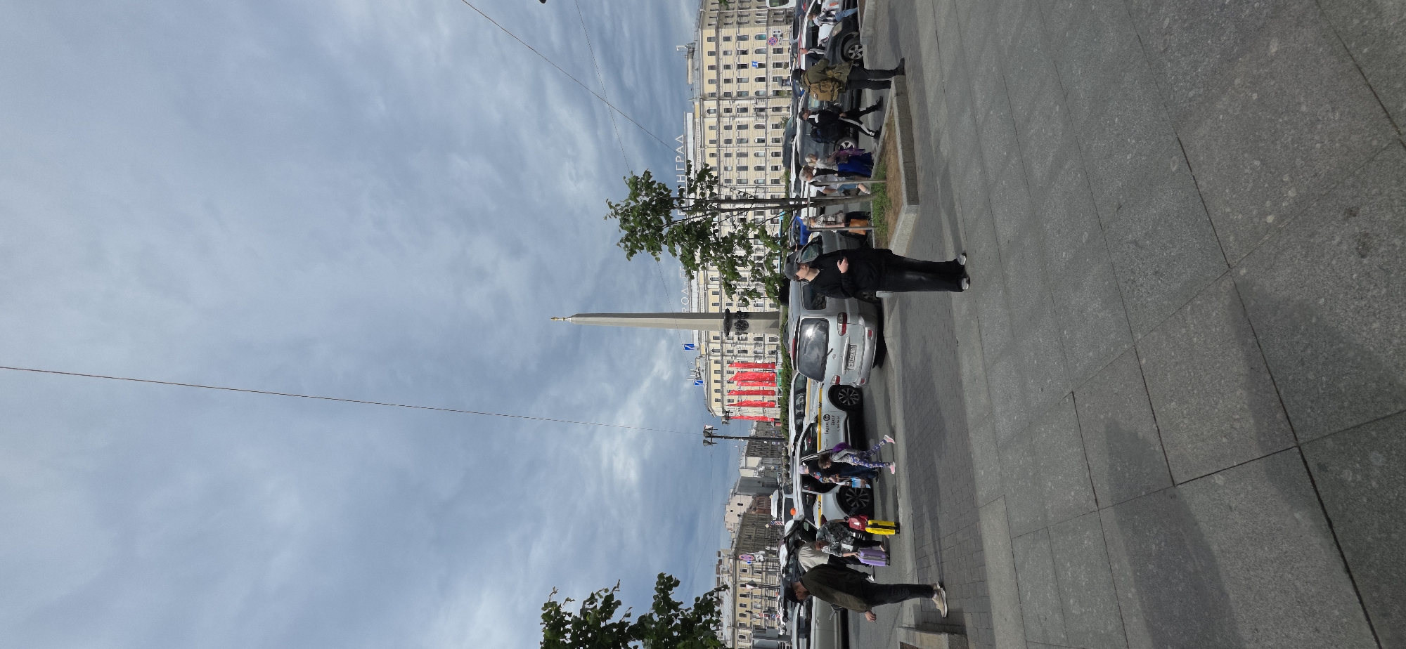 The image captures a street scene with a cloudy sky. In the foreground, a person in a black jacket and pants stands on a paved sidewalk. Behind them, a line of parked cars, including a white minivan and several yellow taxis, are visible. Further in the background, several people are seen standing near the cars, some appearing to be a family with children. A tall, slender monument or obelisk rises above the buildings in the distance, with red banners draped across a nearby structure. The architecture of the buildings suggests an urban environment. There is some visible text on a building in the background, partially obscured but appearing to be in Cyrillic. The scene appears to be set during the daytime.