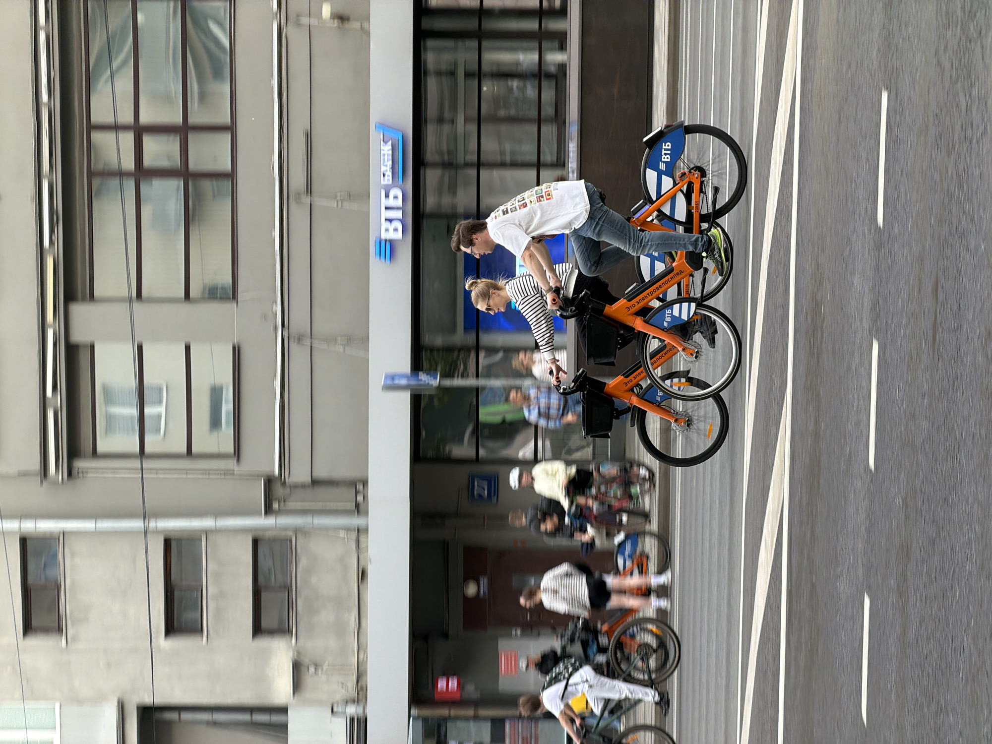 A street scene in Moscow, Russia shows a couple riding orange bicycles on a marked bike path alongside a building. The man in front is wearing a white t-shirt with prints and jeans, and the woman behind him is wearing a striped shirt and dark bottoms. Both appear to be in their 20s or 30s. Several other people on bikes are visible behind them, moving in the same direction. A bank sign with the Cyrillic lettering "ВТБ БАНК" is visible on the building. The street has white lines indicating lanes. The lighting suggests it is daytime. The building's architecture is modern, with multiple windows.