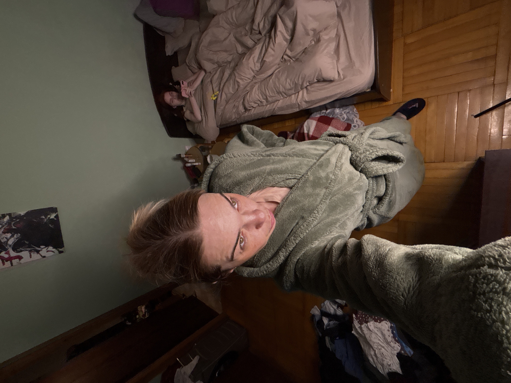 A selfie taken from a high angle shows a person in a green robe. In the background is a bed with another person lying in it. Clothes are scattered on the floor near the person in the robe. The location is a bedroom.
