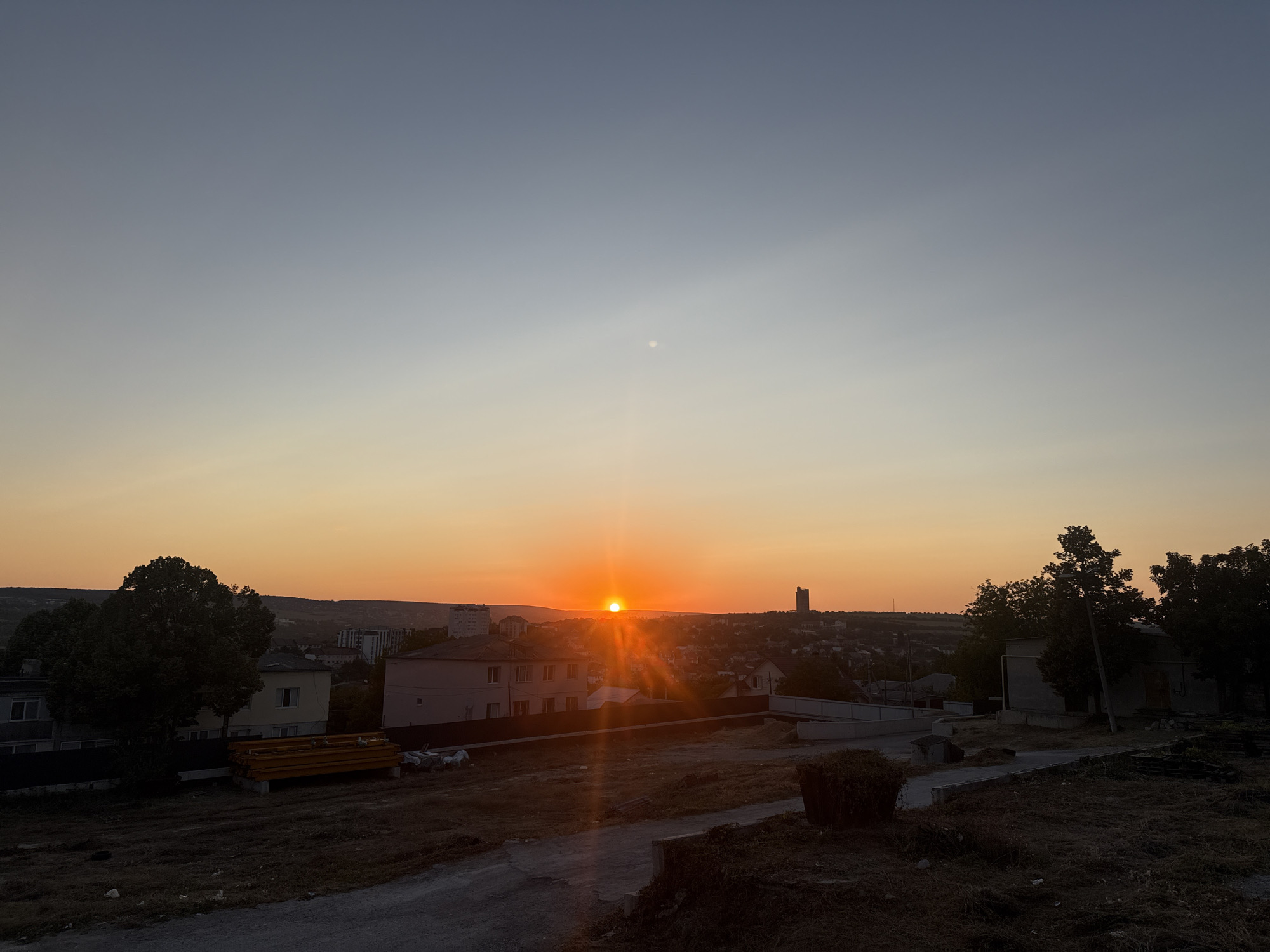 The image depicts a sunset over the city of Ialoveni, Moldova. The sun is a bright orange orb low on the horizon, casting long rays of light across the sky. The sky transitions from a pale blue at the top to a warm orange near the horizon.
In the foreground, there is a dirt path with dry, sparse vegetation. To the left, a large, dark silhouette of a tree stands prominently. In the middle ground, several buildings are visible, including a light-colored house with windows and a darker-pitched roof, and another building with a pinkish facade. A yellow structure, possibly construction materials, is also present.
The background features a cityscape with more buildings, some appearing to be residential. A tall, slender tower-like structure is visible in the distance on the right side of the frame. The landscape behind the city is hilly and appears to be covered in vegetation. The overall scene is one of a quiet, rural or semi-urban environment at dusk. There are no people visible in the image.