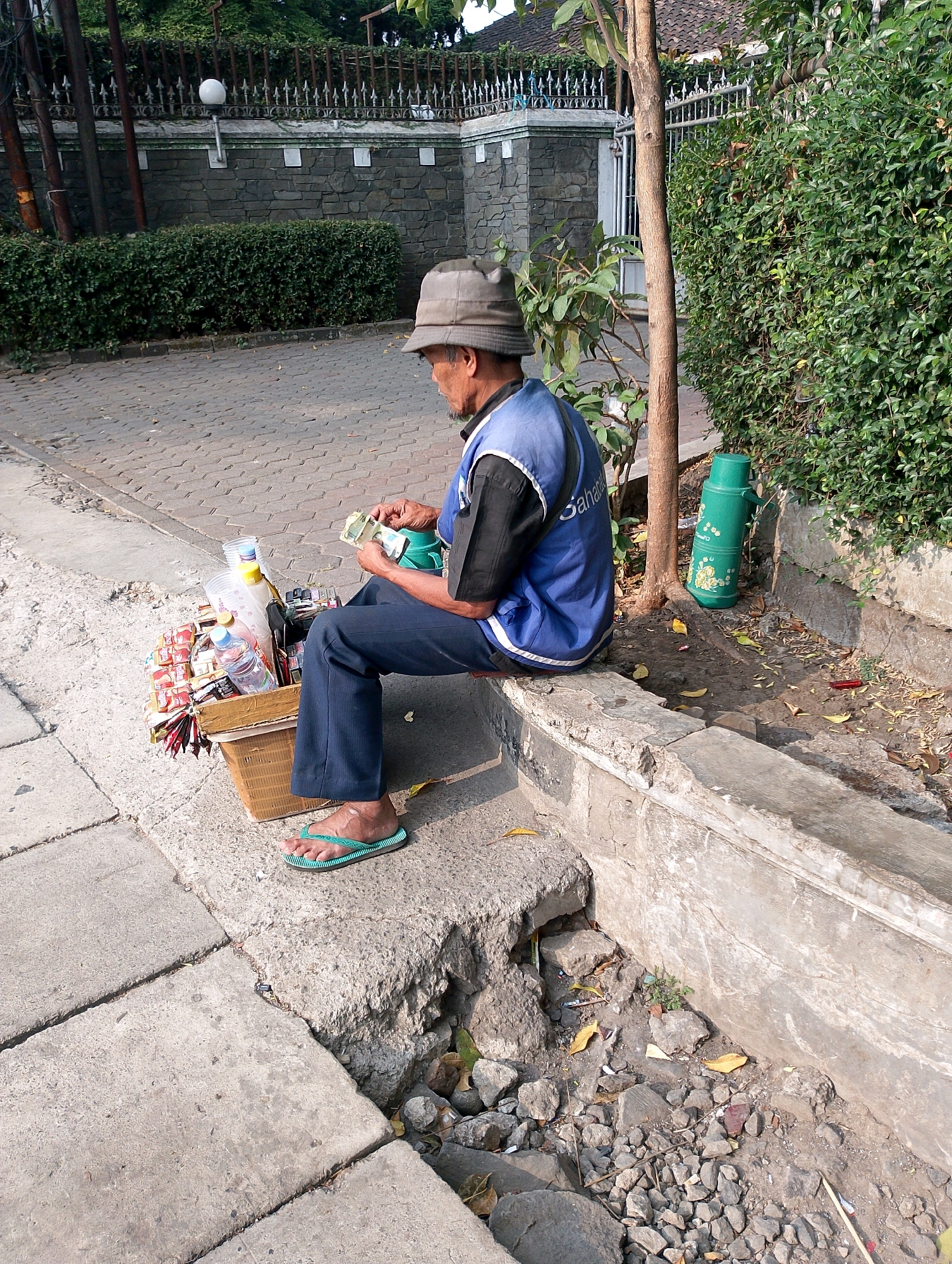 A man wearing a light-colored hat and a blue vest over a dark shirt sits on a concrete ledge, counting money. He is wearing dark pants and green flip-flops. Beside him is a woven basket filled with bottled drinks, snacks, and plastic cups. Behind him, a green thermos with a floral pattern sits near a tree. The man is in an outdoor urban setting, with a stone wall and lush green bushes visible in the background. The ground is paved with interlocking bricks and concrete slabs, with scattered stones and fallen leaves. This scene is located on a street in Bandung City, Indonesia.