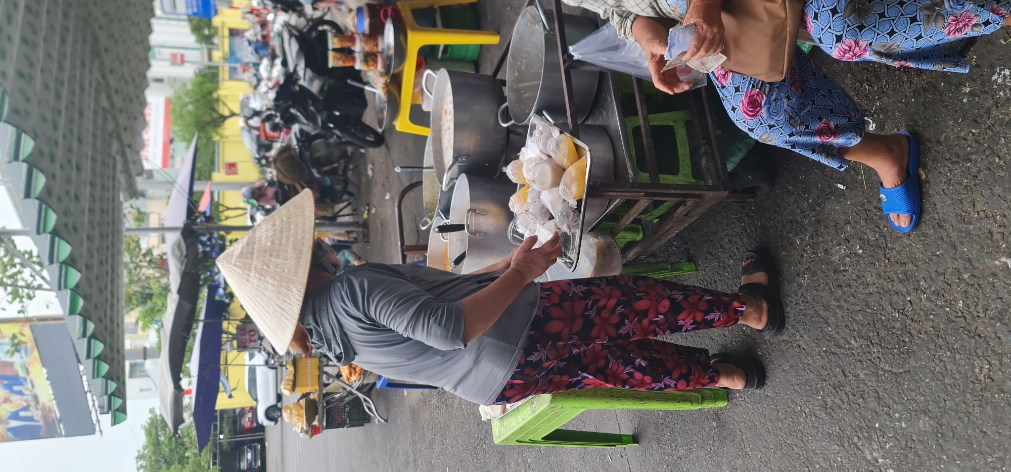 A vendor wearing a traditional conical hat and a gray hooded long-sleeve shirt and flower pants is standing behind a metal pushcart laden with large metal pots and a metal tray filled with clear plastic bags of food. She appears to be handing a bag to a customer. The customer, seated on a plastic green chair to the right, is wearing blue floral print pants and blue sandals. She is holding money. The scene is a street in a city, likely Đà Nẵng, Vietnam, based on the style of the hat and vendor's clothing. In the background, several parked motorcycles and vehicles can be seen, as well as buildings and awnings.