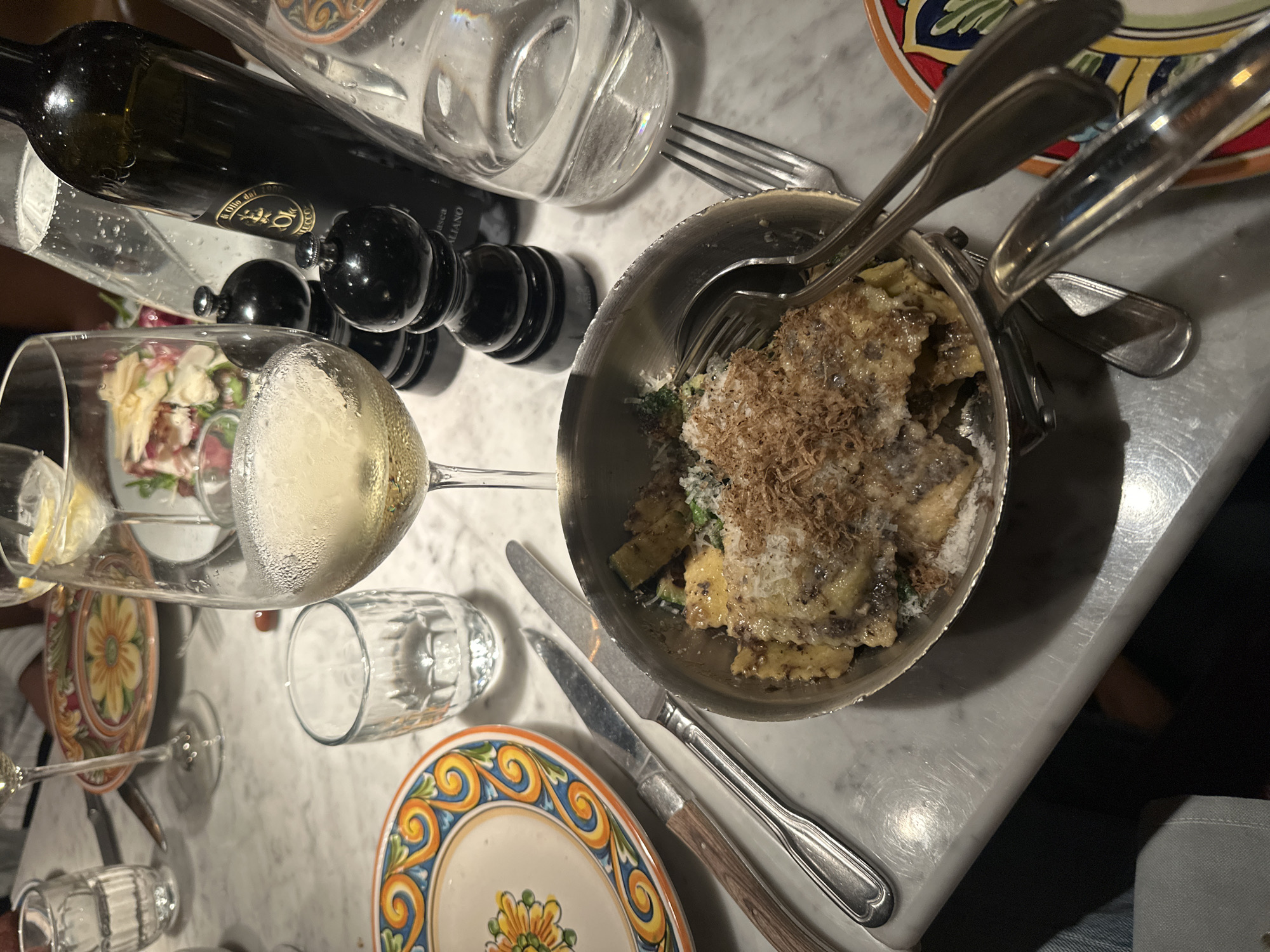 An overhead shot of a table setting in Copenhagen, Denmark, displays a meal in progress. The table, made of marble, holds several plates, cutlery, glassware, and a bottle of olive oil. In the foreground, a silver bowl contains a dish of pasta, topped with grated cheese and what appears to be truffles. Two forks are placed inside the bowl. To the left of the bowl, a knife with a wooden handle rests on a decorative plate. A wine glass filled with white wine is positioned behind the bowl, and a smaller glass filled with water sits to its left. Two black salt and pepper grinders are visible behind the wine glass. A bottle of olive oil stands to the right of the grinders. In the background, a partially visible salad can be seen on another plate. The overall scene suggests a dining experience at a restaurant.