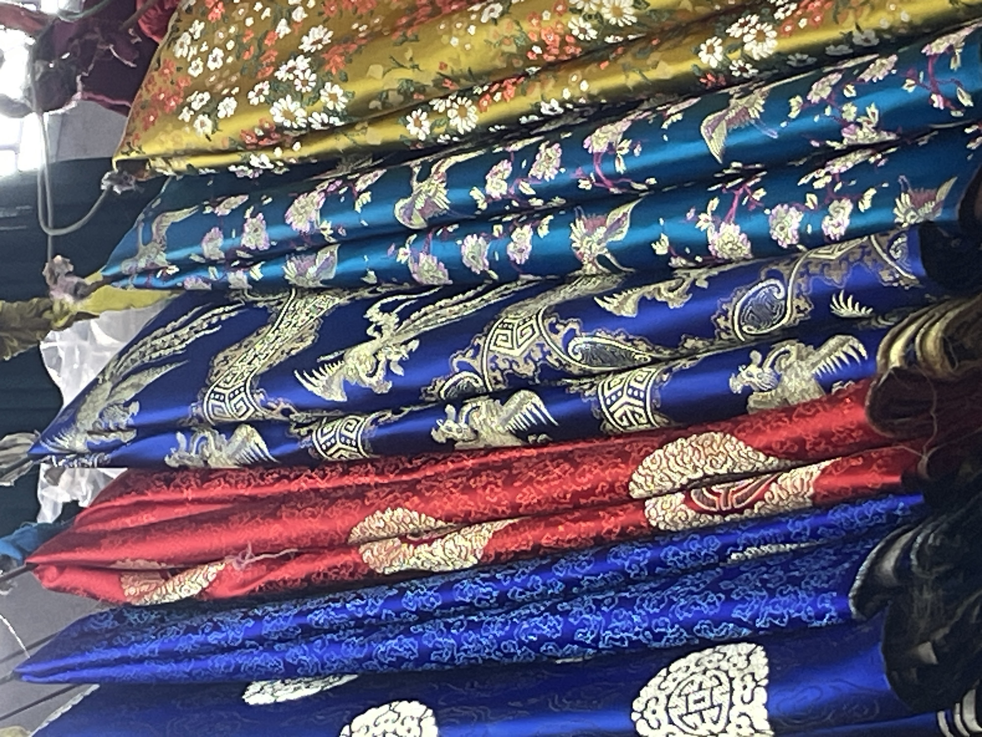 A close-up, eye-level, medium shot captures a vibrant display of four richly patterned silk fabrics hanging vertically and slightly angled from a rail. The fabrics are arranged from left to right in the following order: a royal blue fabric adorned with a white circular emblem and intricate blue swirling patterns, a bright red fabric with a white and gold ornate pattern, a deep blue fabric featuring gold phoenixes and geometric designs, and a teal fabric with a floral and bird motif in pink, purple, and white. To the far right, a section of gold fabric patterned with small white and pink flowers is visible. The fabrics are draped and folded, revealing their lustrous sheen and detailed embroidery. The background is dark and out of focus, suggesting an indoor setting, possibly a market stall or textile shop. The lighting is moderate, highlighting the textures and colors of the silks. No people are visible in the frame, and no specific location within Nha Trang, Vietnam can be determined from the image alone; it is a generic depiction of high-quality textiles.
