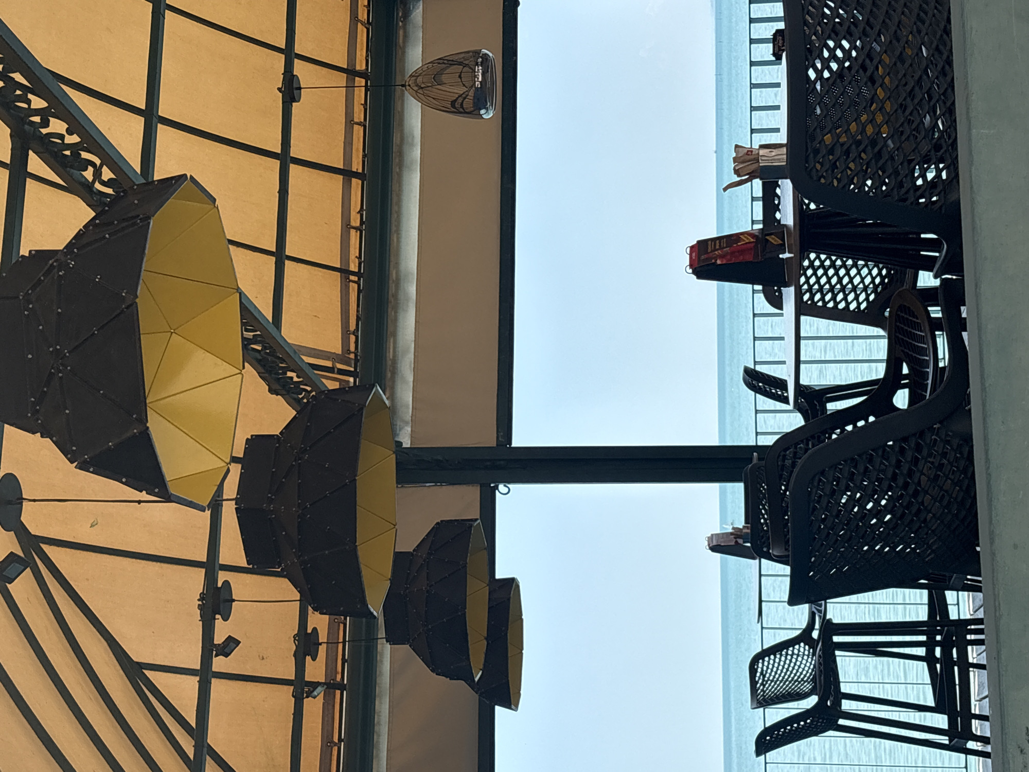 The image depicts an outdoor seating area with a view of the ocean. On the left side of the image, there are several large, conical light fixtures with black exteriors and yellow interiors, suspended from a tan canopy supported by a dark metal frame. The canopy appears to be made of fabric and is stretched across the frame.
On the right side of the image, there are dark, plastic chairs and tables arranged in front of a railing. Some of the tables have items on them, including what appear to be small boxes or packages and possibly food items. The chairs have a woven pattern.
In the background, a body of water, likely the ocean, is visible. The sky is a pale blue, suggesting daytime, possibly late morning or early afternoon. The lighting is bright, indicating fair weather.
The overall scene appears to be a cafe, restaurant, or a similar establishment with outdoor seating, possibly near a waterfront. There are no people visible in the image. The perspective is from below, looking upwards and across the scene, with the image rotated 90 degrees counter-clockwise. The composition suggests a modern, possibly tropical or coastal, setting. There are some small, dark, square lights attached to the metal framework supporting the canopy.