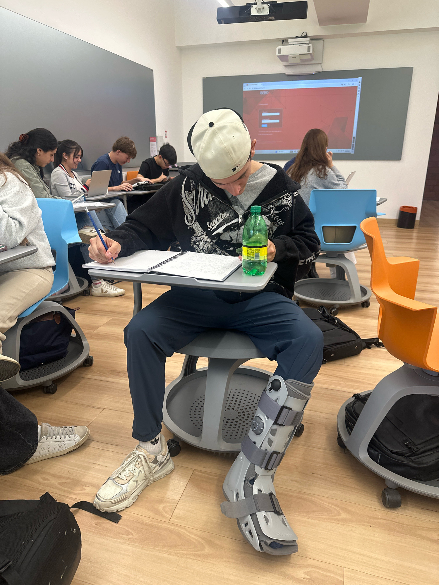 In a classroom in Mexico City, Mexico, several students are seated at desks. In the foreground, a young man wearing a black hooded sweatshirt, dark pants, and a white baseball cap is focused on writing in a notebook with a pencil. He has a green bottle of water next to his notebook and is wearing a walking boot on his right foot. Other students are visible in the background, some using laptops and others engaged in note-taking. A projector is mounted on the ceiling, displaying an image on a screen at the front of the classroom. The room has modern, rolling chairs and desks, and the flooring is a light-colored wood.