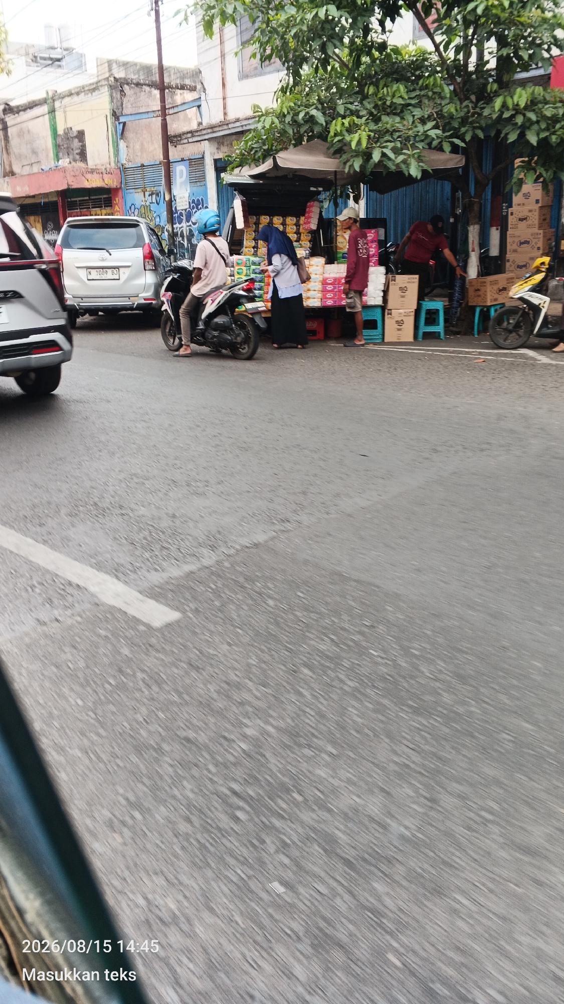 This is a medium shot, taken from a moving vehicle, of a street scene in Malang, Indonesia. A silver car with license plate N 1089 BJ is parked on the left side of the road. In front of the car, a person wearing a light-colored shirt and blue helmet is sitting on a black motorcycle. To the right of the motorcycle, a woman wearing a dark hijab and a light-colored shirt is standing next to a roadside stall. The stall has an umbrella canopy and displays stacks of products, appearing to be toiletries or packaged food items. Two men are also visible near the stall. One man, wearing a maroon shirt, is standing and looking towards the stall. Another man, wearing a dark t-shirt and baseball cap, is partially visible behind the stall, reaching for something. There are several blue plastic stools and stacks of cardboard boxes around the stall. A yellow motorcycle is parked to the right of the stall. The road surface is asphalt with a white line marking a lane. The background shows buildings with weathered facades and trees. The image has a timestamp at the bottom left indicating "2026/08/15 14:45" and "Masukkan teks".