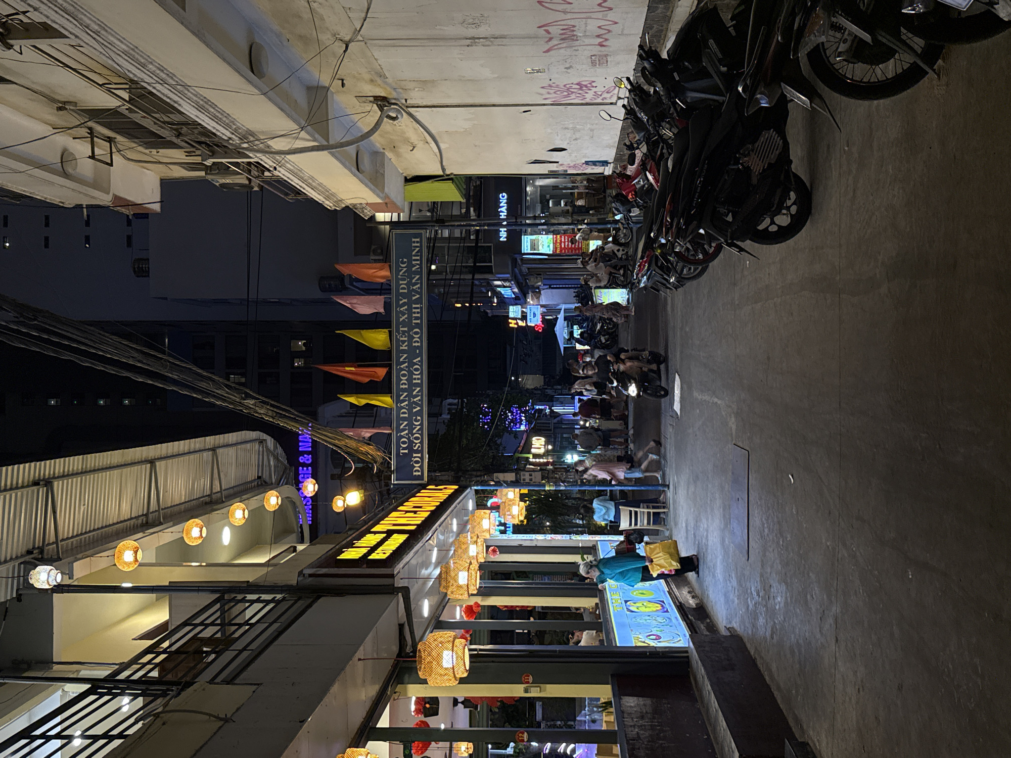 The image captures a lively night street scene in what appears to be an urban area, likely Nha Trang Ward, Vietnam. The setting is illuminated by a multitude of artificial lights, including glowing lanterns, bright shop signs, and streetlights, indicating it is nighttime.
Numerous motorbikes are parked densely along one side of the street, a common sight in Vietnamese cities. Buildings with various awnings and corrugated metal roofs line the thoroughfare. On the left, several decorative woven lanterns hang, casting a warm, inviting glow, while more elaborate lanterns extend further down the street on the right.
Several people are visible, engaged in various activities. Some are seated at outdoor tables, likely dining or socializing at street-side eateries. One individual in a light blue jacket stands near a brightly lit display, holding yellow bags, possibly shopping. Another person is seen on a motorbike further down the street. Colorful, triangular flags are strung across or from buildings, adding a festive touch.
Visible text on signs includes:
- A prominent vertical sign reads: "TOÀN DÂN ĐOÀN KẾT XÂY DỰNG ĐỜI SỐNG VĂN HÓA - ĐÔ THỊ VĂN MINH" (translates to "The entire population unites to build a cultural lifestyle - civilized urban area").
- Another sign clearly states "NH HÀNG" (Restaurant).
- Fragments like "MAS&GE & NA" (likely "Massage & Nail") and "MIO" are also visible. Graffiti is scrawled on one of the walls.
The scene conveys a bustling and active atmosphere, typical of an evening in a vibrant Vietnamese city.