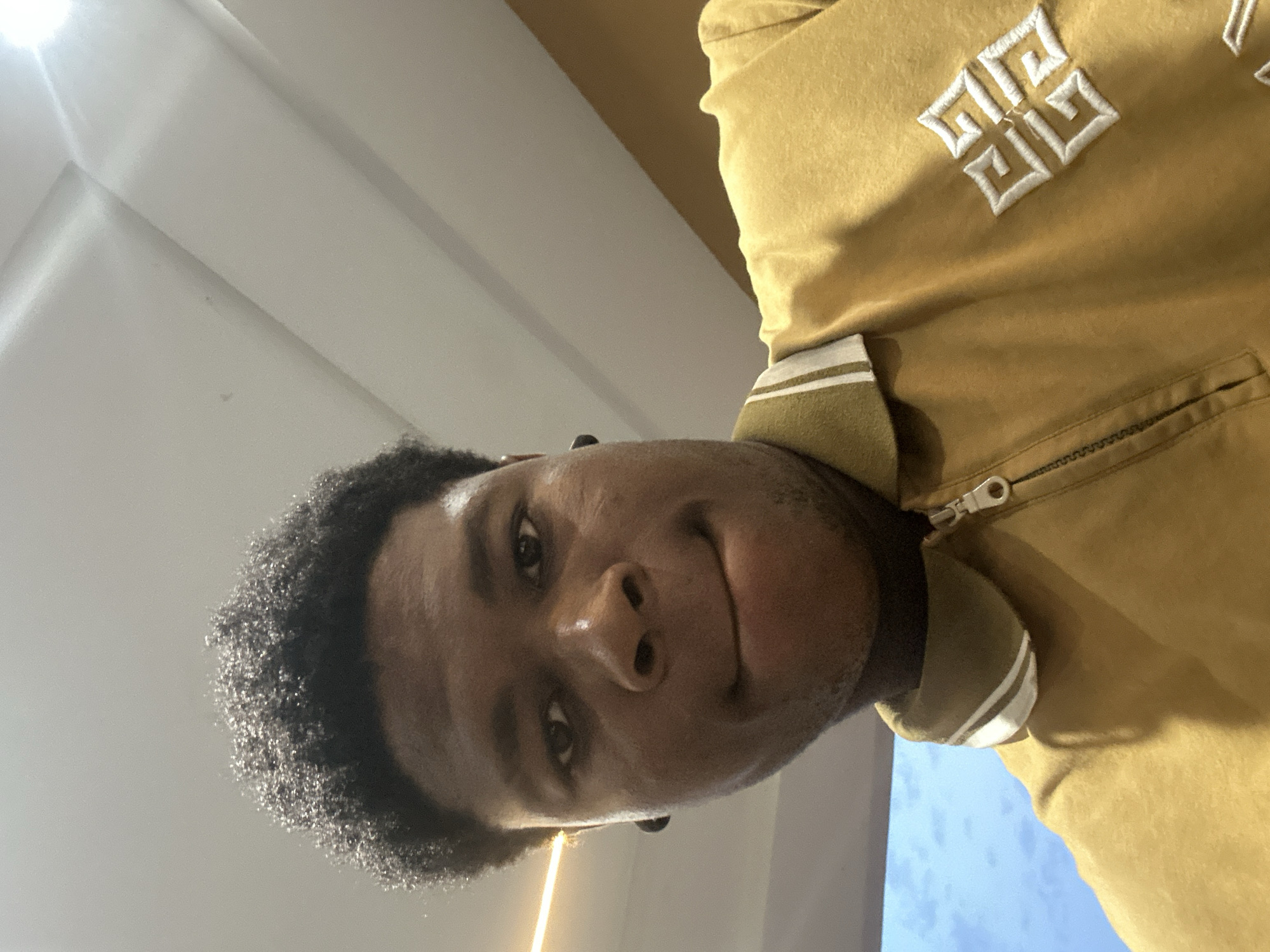 The image is a close-up, low-angle selfie of a young Black male. He has dark, curly hair and is wearing a yellow polo shirt with white trim on the collar and a zipper. There is a white embroidered logo on the right side of his shirt. He is biting his lower lip and looking directly at the camera. The background appears to be an indoor environment with a white ceiling and what looks like a light fixture on the left side. The overall lighting is bright, and the image quality is good. No specific location within Ihiagwa, Nigeria, can be determined from the visual information provided in the image.