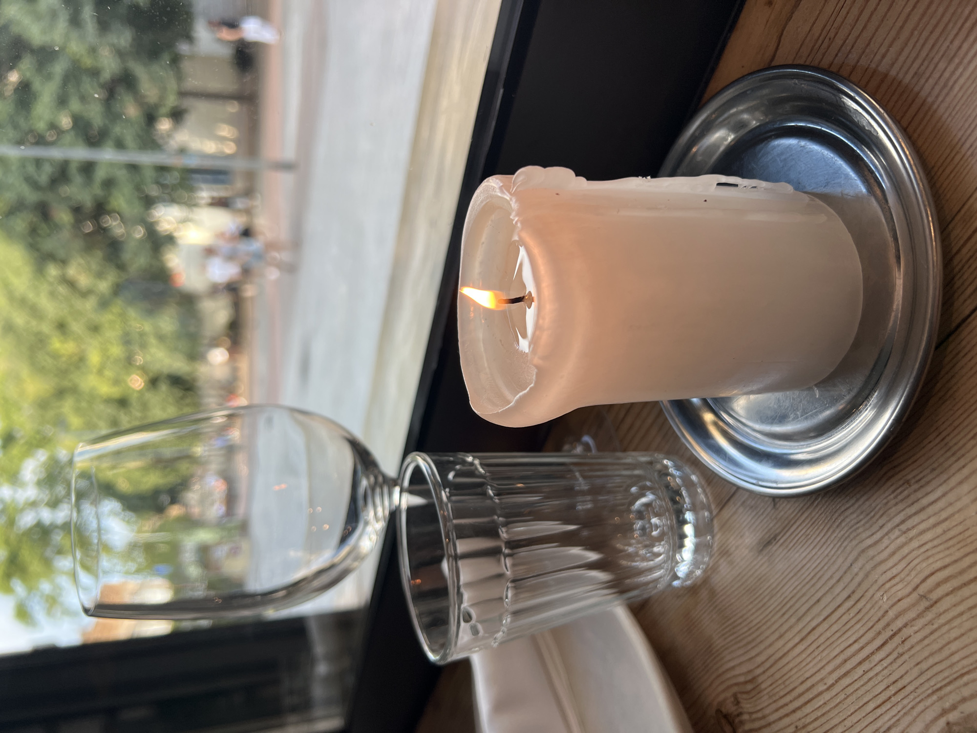 A lit white candle sits on a small silver saucer on a wooden table, next to two empty glasses. One is a wine glass, and the other is a small, fluted glass. In the background, through a window, trees and people are visible on a street. The scene is likely a cafe or restaurant in Barcelona, Spain, given the Mediterranean foliage and pedestrian activity.