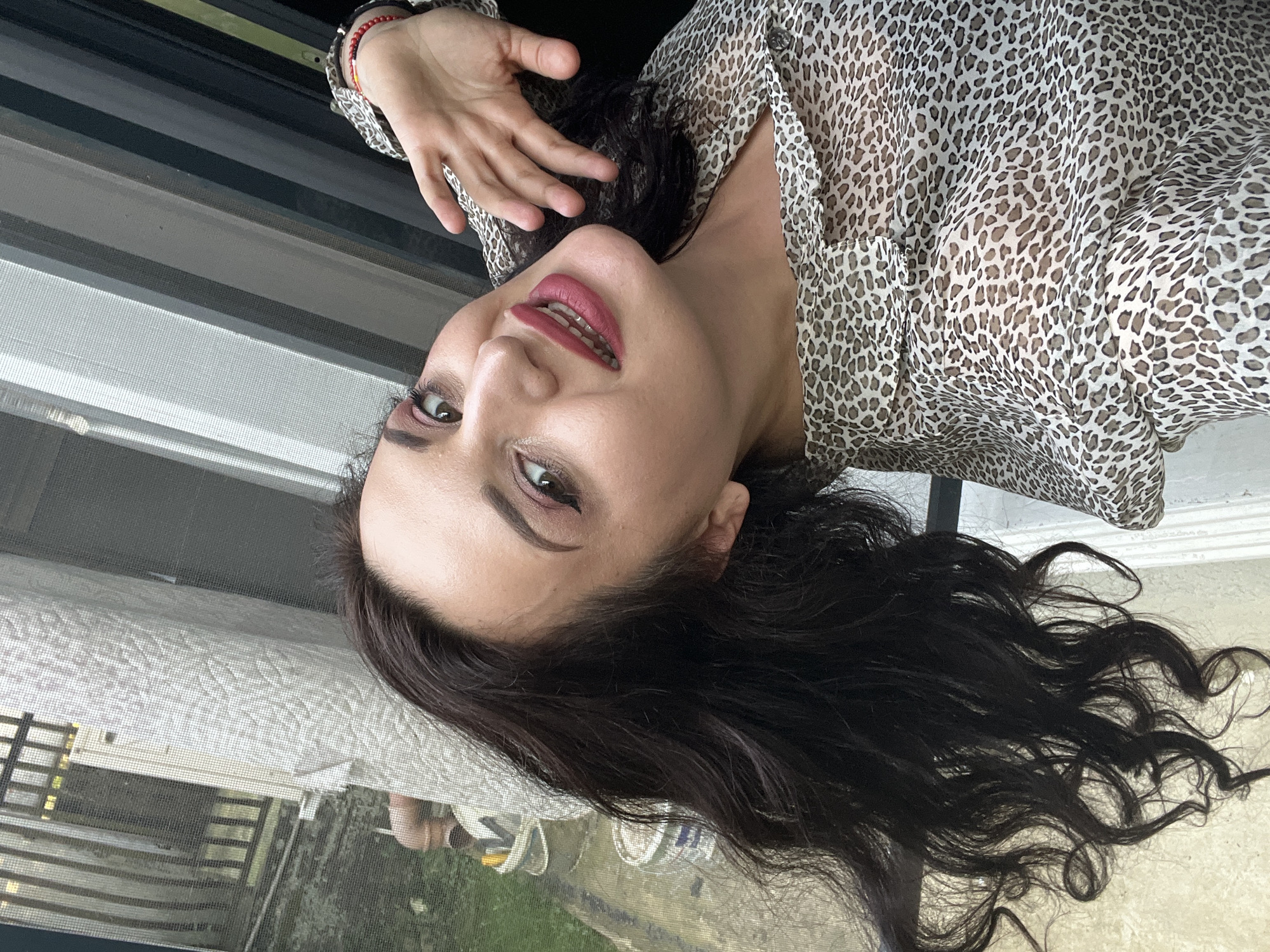 This is a close-up, angled selfie-style portrait of a woman. The woman has long, dark, wavy hair and fair skin. Her facial features include defined eyebrows, dark eyes, and a wide smile revealing visible dental braces on her upper teeth. She is wearing winged eyeliner and pink-red eyeshadow and lipstick. Her attire consists of a translucent, light-colored blouse with a distinct leopard print pattern of irregular brown/black spots on an off-white background. A red beaded bracelet is visible on her right wrist, which is raised with fingers splayed near her face.
The photograph is taken indoors, looking outwards through a fine mesh screen that covers the entire left and central background, creating a slightly blurred, textured overlay. To the left, visible through the screen, is an outdoor area featuring green ground, two white buckets, and a section of a dark, pipe-like structure. Further back, a wooden slatted fence or barrier is visible. To the right of the woman, beyond the mesh screen, a set of vertical striped window blinds in alternating dark and light gray are partially visible. The lighting is natural and diffuse, suggesting an outdoor light source. The location is an unspecified residential or building setting in Unknown, Unknown.