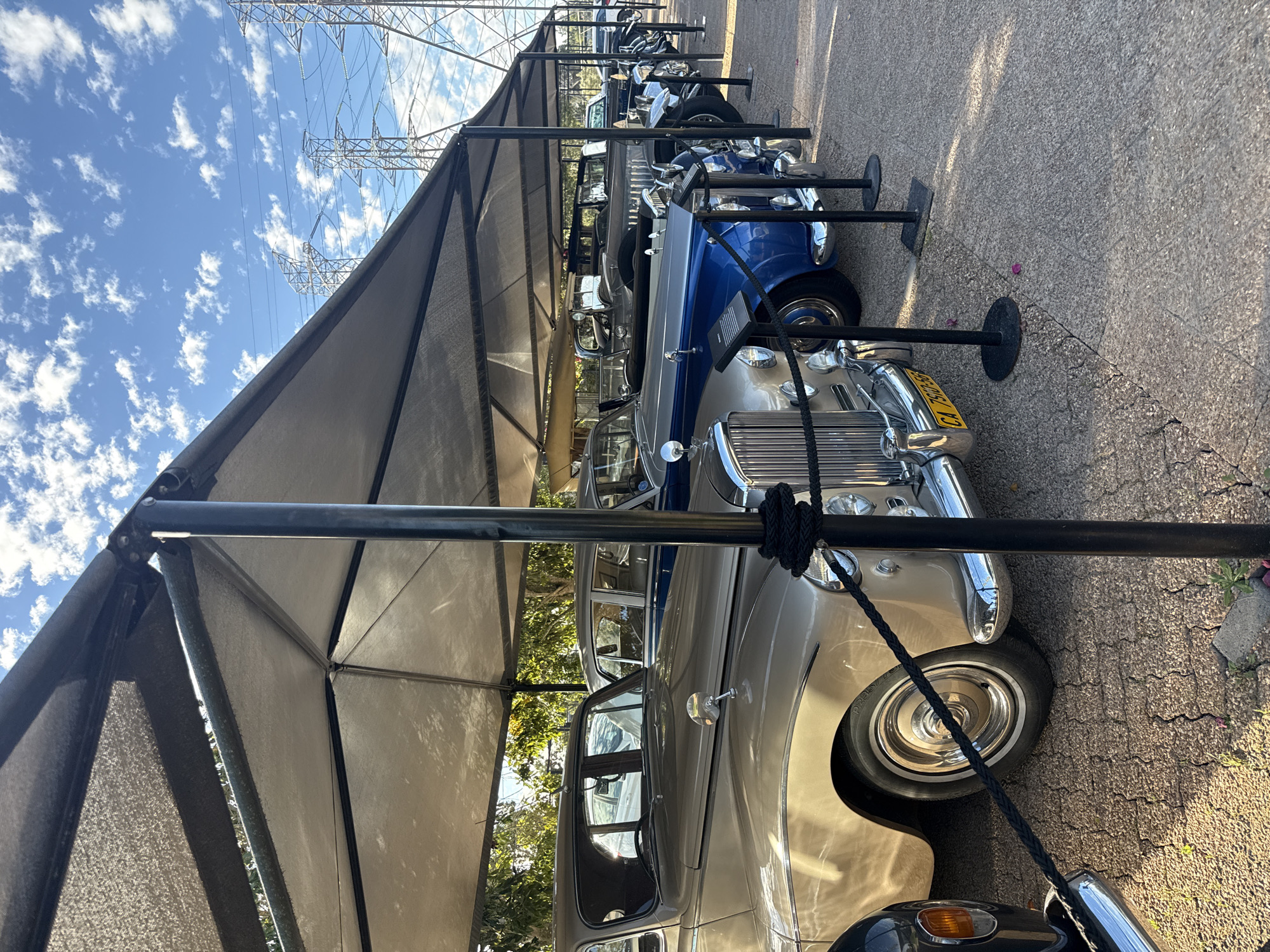 The image depicts a row of vintage cars parked under a shaded structure in Stellenbosch Nu, South Africa. The cars are lined up on a paved surface with black metal posts and ropes delineating the display area. Overhead, high-voltage power lines are visible against a blue sky with scattered clouds. The vehicles are a mix of colors and styles, primarily older models. The light suggests it is a sunny day. There are no people in the immediate view of the frame.
