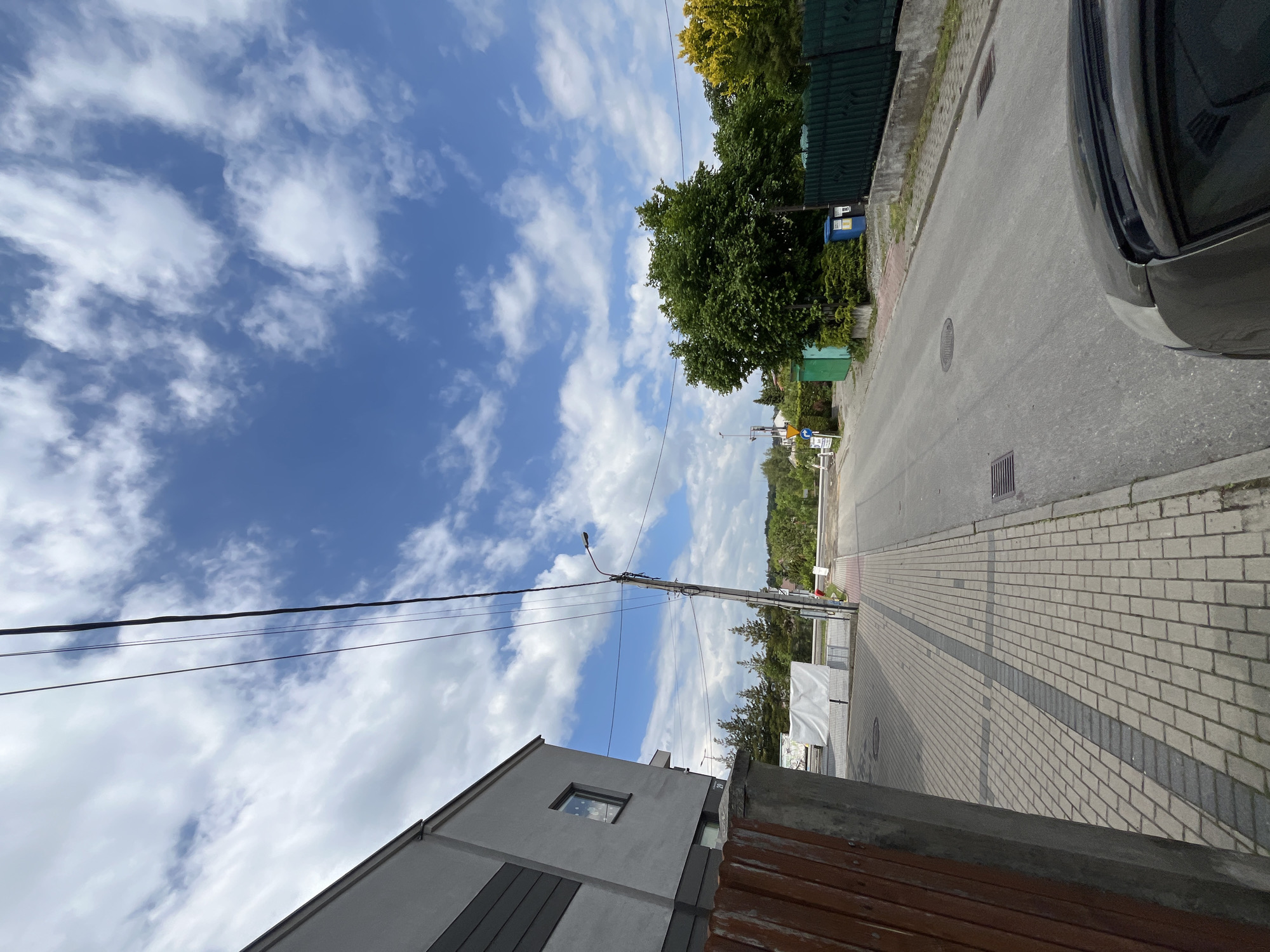 The image captures a street scene in an unidentified city. The view is oriented upwards, showing a street with a paved sidewalk on the right and a road on the left. A gray car partially obscures the top right corner of the frame.
On the left side of the street, there's a modern gray building with a single visible window. Utility poles with wires extend across the sky, which is predominantly blue with scattered white clouds. On the right side of the street, greenery including trees and fences lines the road. Further down the street, there's more vegetation and a glimpse of buildings in the distance. Street signs are also visible in the distance. The sky is bright and indicates a sunny day.