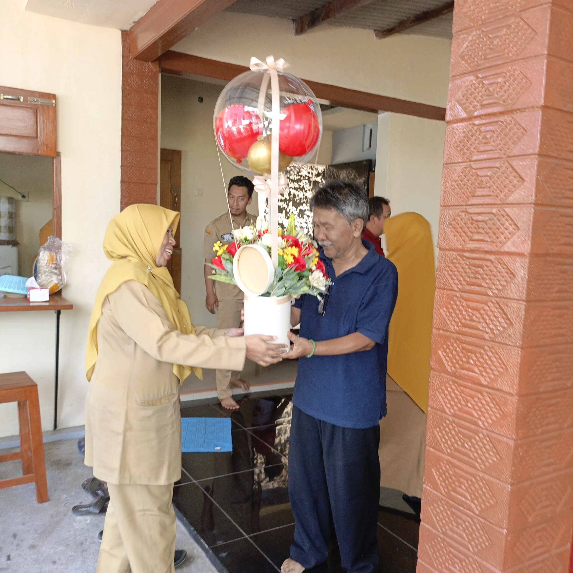 The image shows a man and a woman standing in what appears to be the entrance of a building. The woman, on the left, is wearing a light brown uniform and a yellow hijab. She is smiling and holding a white cylindrical container filled with flowers. The man, on the right, is wearing a dark blue polo shirt and black pants. He is looking at the flowers and appears to be receiving them. Behind them, other people are visible, some in uniforms. A large clear balloon with red and gold spheres inside is suspended by ribbons above the flowers. The environment suggests a semi-outdoor setting with tiled floors and a pillar with a textured orange surface. The building has wooden elements and a metal roof. The presence of uniforms and the formal attire of the flower-giver suggest this might be an official event or ceremony. The location is identified as Salatiga, Indonesia.