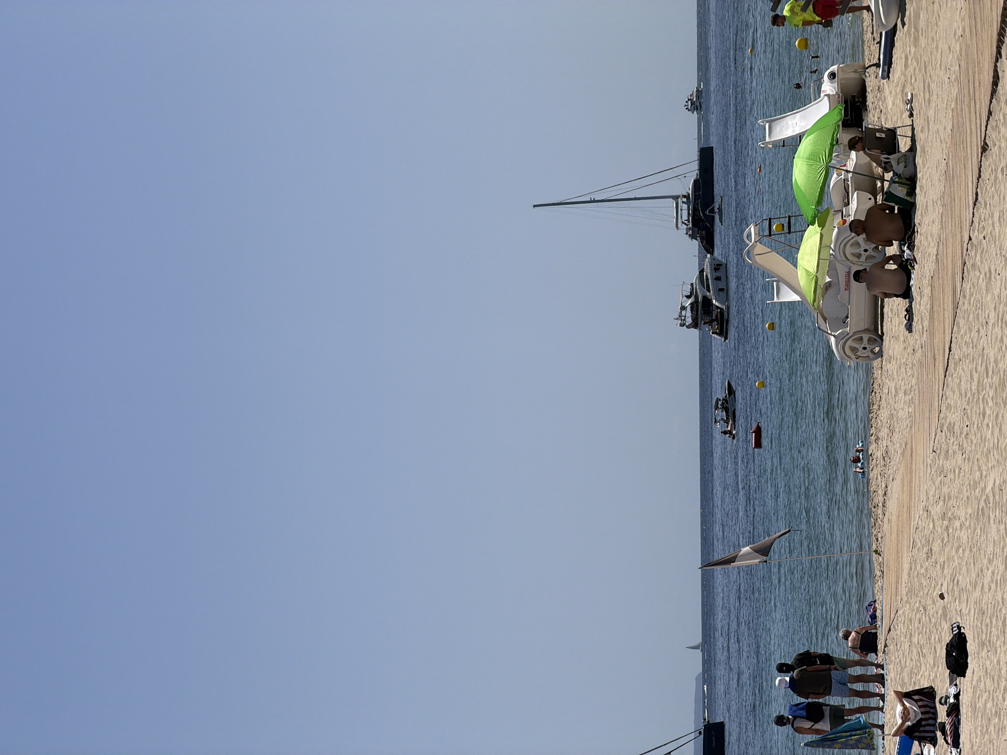 The image depicts a sunny day at Palmanova beach in Spain. The foreground shows a sandy beach with wooden pathways. Several people are relaxing on the sand, some under beach umbrellas. A few white pedal boats with slides are parked near the water's edge. In the background, the blue sea is dotted with various boats, including yachts and smaller vessels. A few people are in the water, and a yellow buoy is visible. The sky is clear and blue.