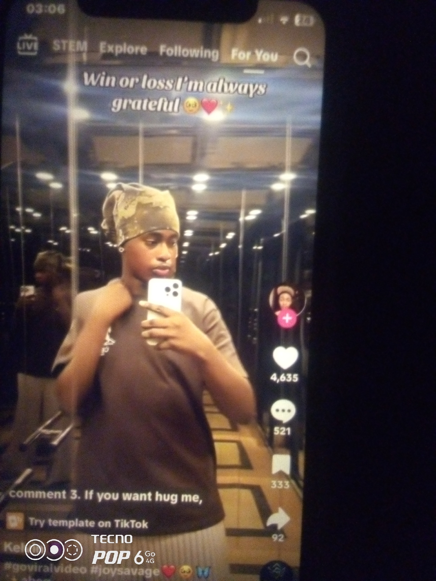 The image displays a selfie taken by a young Black person wearing a bandana and a brown t-shirt, holding a white smartphone. The person is positioned in front of a reflective surface, likely a mirror in an elevator, as indicated by the vertical lines of the elevator doors and the overhead lighting. In the background, another person is visible as a reflection. The screen of the smartphone displays a TikTok interface with text overlaying the video. The visible text reads "Win or loss I'm always grateful 🥺❤️". Below the video, there are interaction icons typical of social media platforms, including a heart with a count of "4,635", a speech bubble with "521", a bookmark with "333", and a share arrow with "92". At the bottom of the screen, a caption appears to start with "comment 3. If you want hug me," followed by "Try template on TikTok" and hashtags like "#goviralvideo" and "#joysavage". The image is dark, suggesting it was taken in low light conditions. There is no discernible information in the image that places the scene specifically in Howayi, Nigeria.